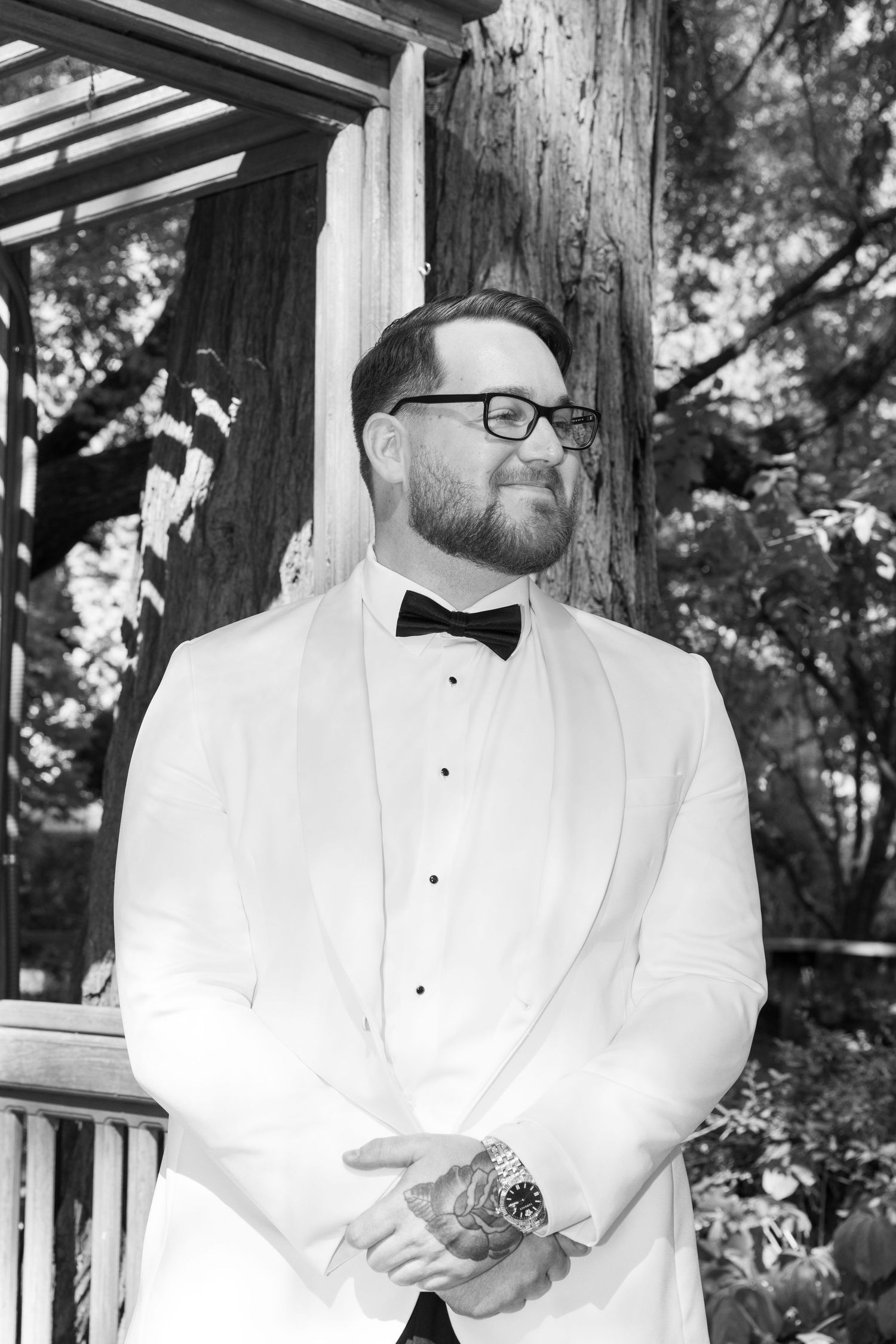 Groom in white tuxedo jacket and bow tie stands on wooden deck surrounded by trees.