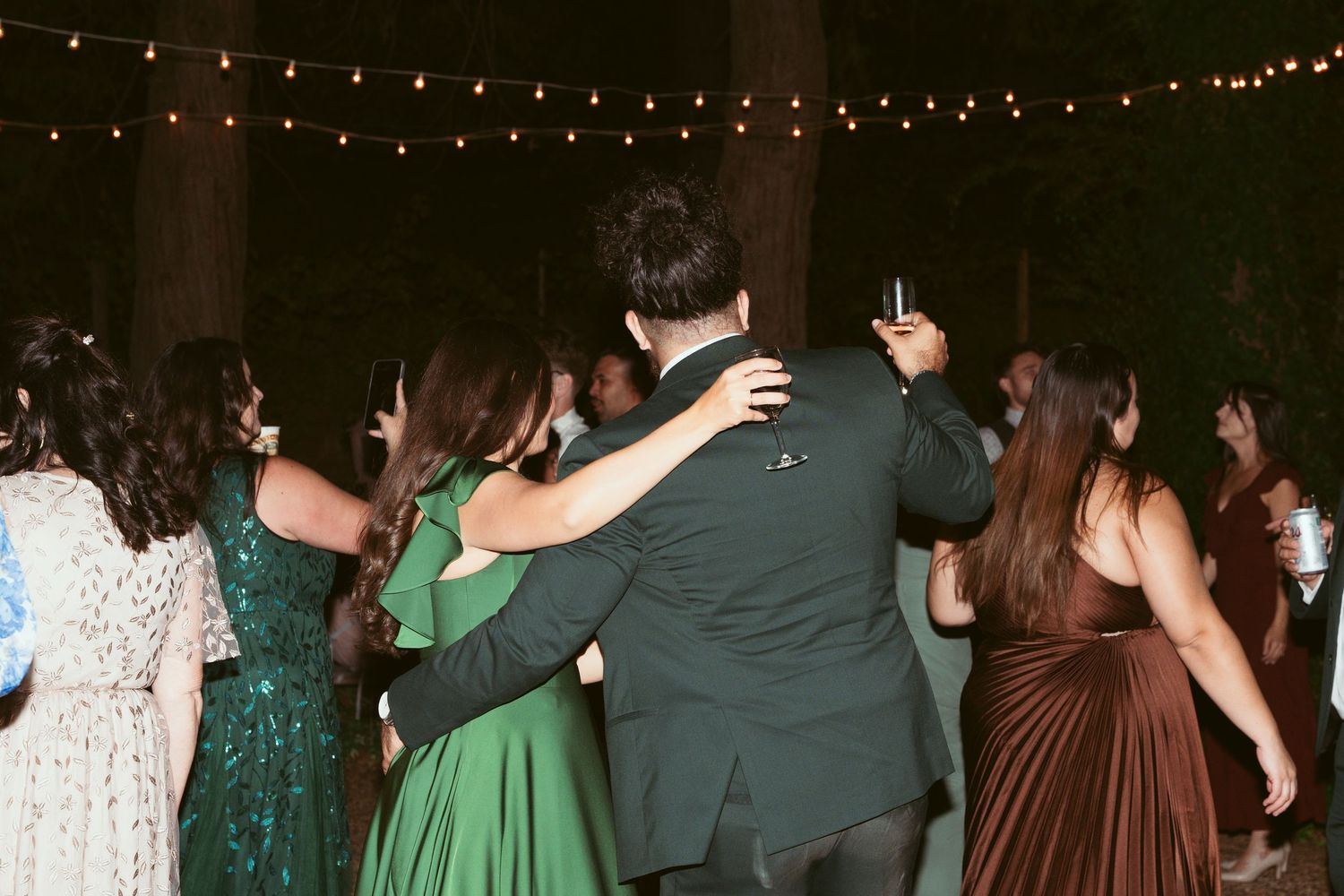 Wedding guests dancing and celebrating under string lights at an outdoor evening reception.