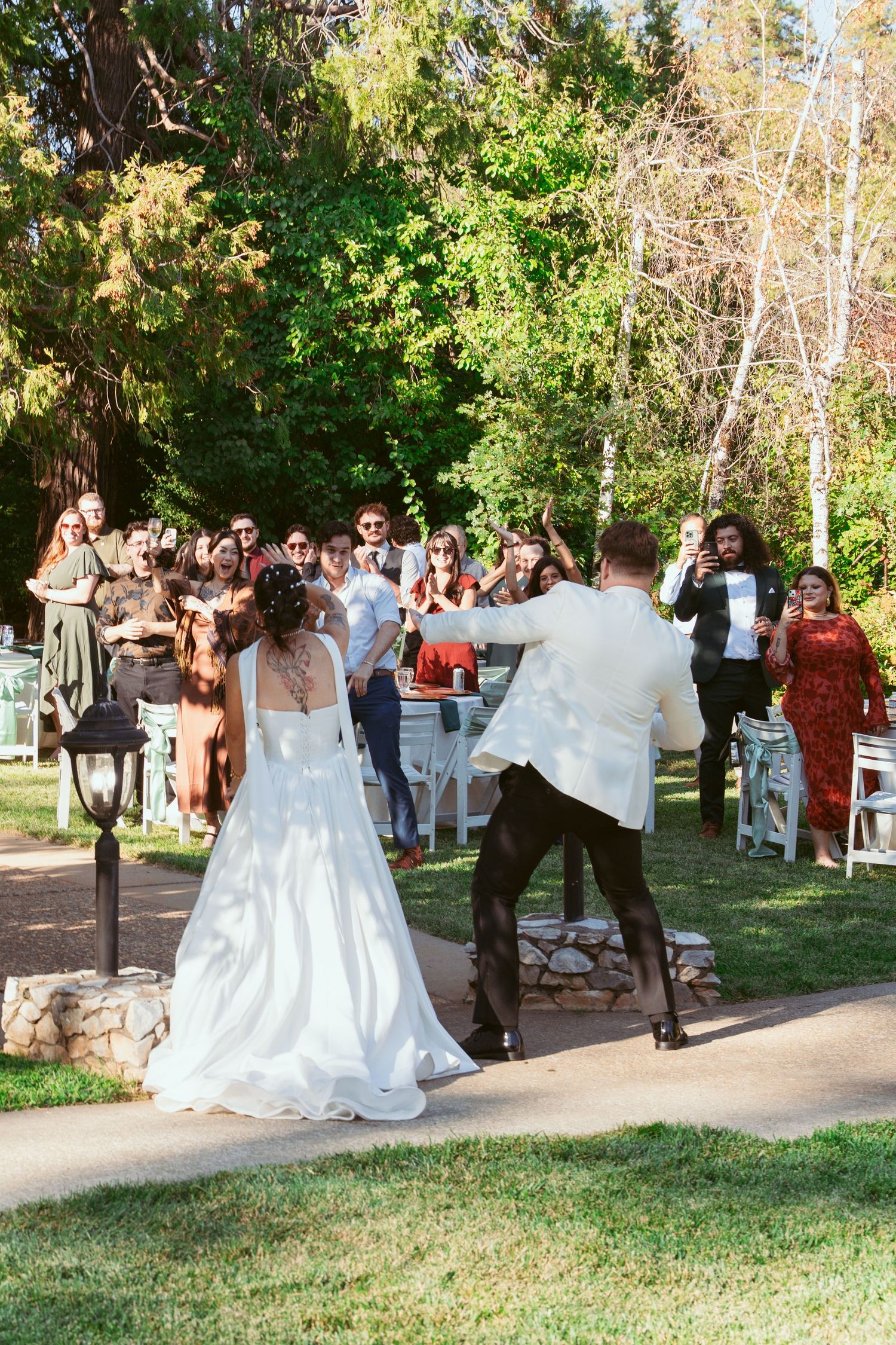 Wedding celebration taking place outdoors with guests gathered among trees and greenery.