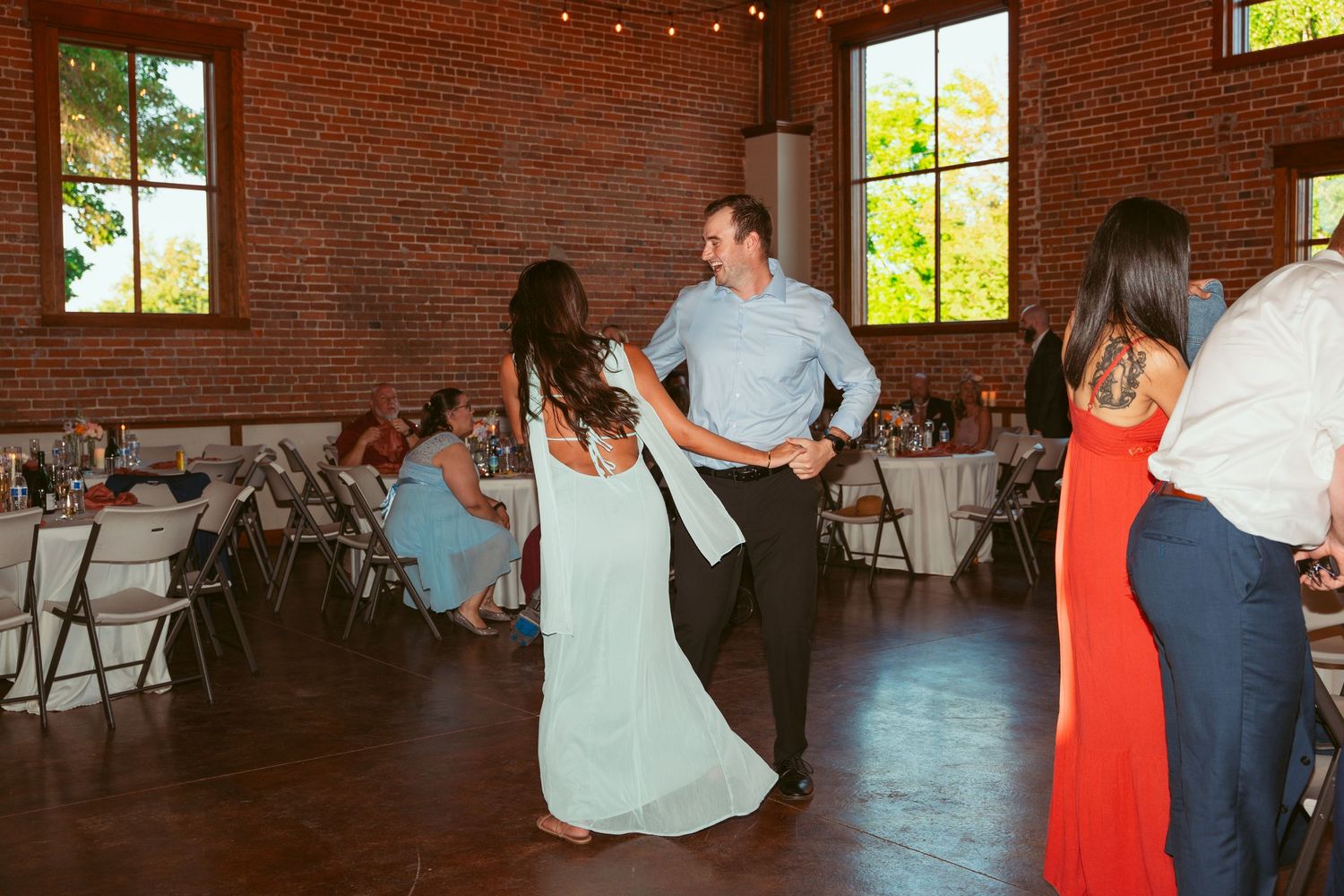 Couples dancing together at a wedding reception in a brick-walled venue with large windows.
