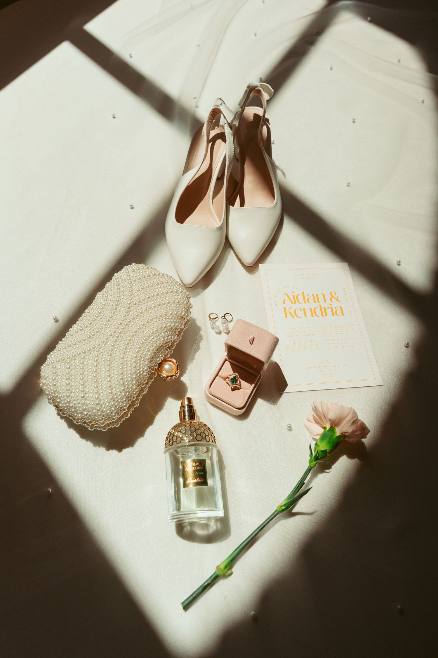 Wedding details with white heels, pearl clutch, perfume bottle and pink flower in dramatic lighting.
