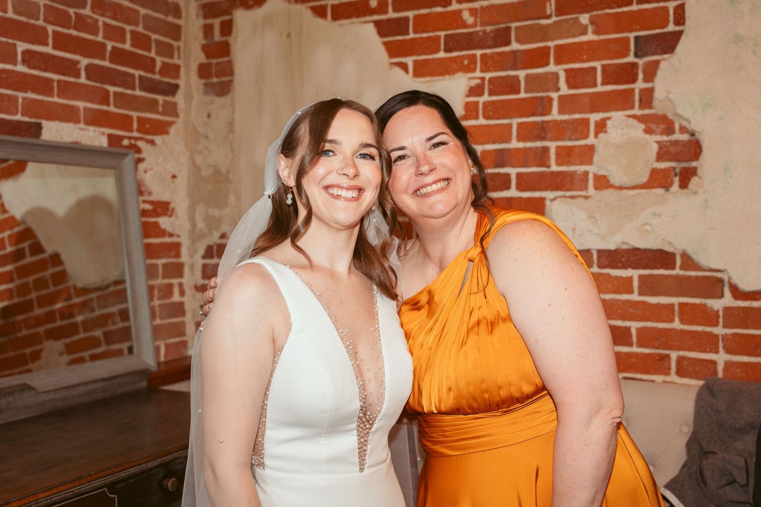 Two people in formal attire share a joyful moment in front of a rustic brick wall.