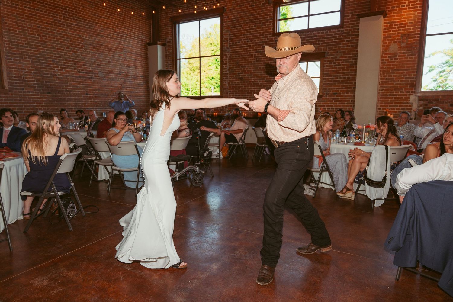 Dancers performing energetic moves in rustic brick reception space with string lights.