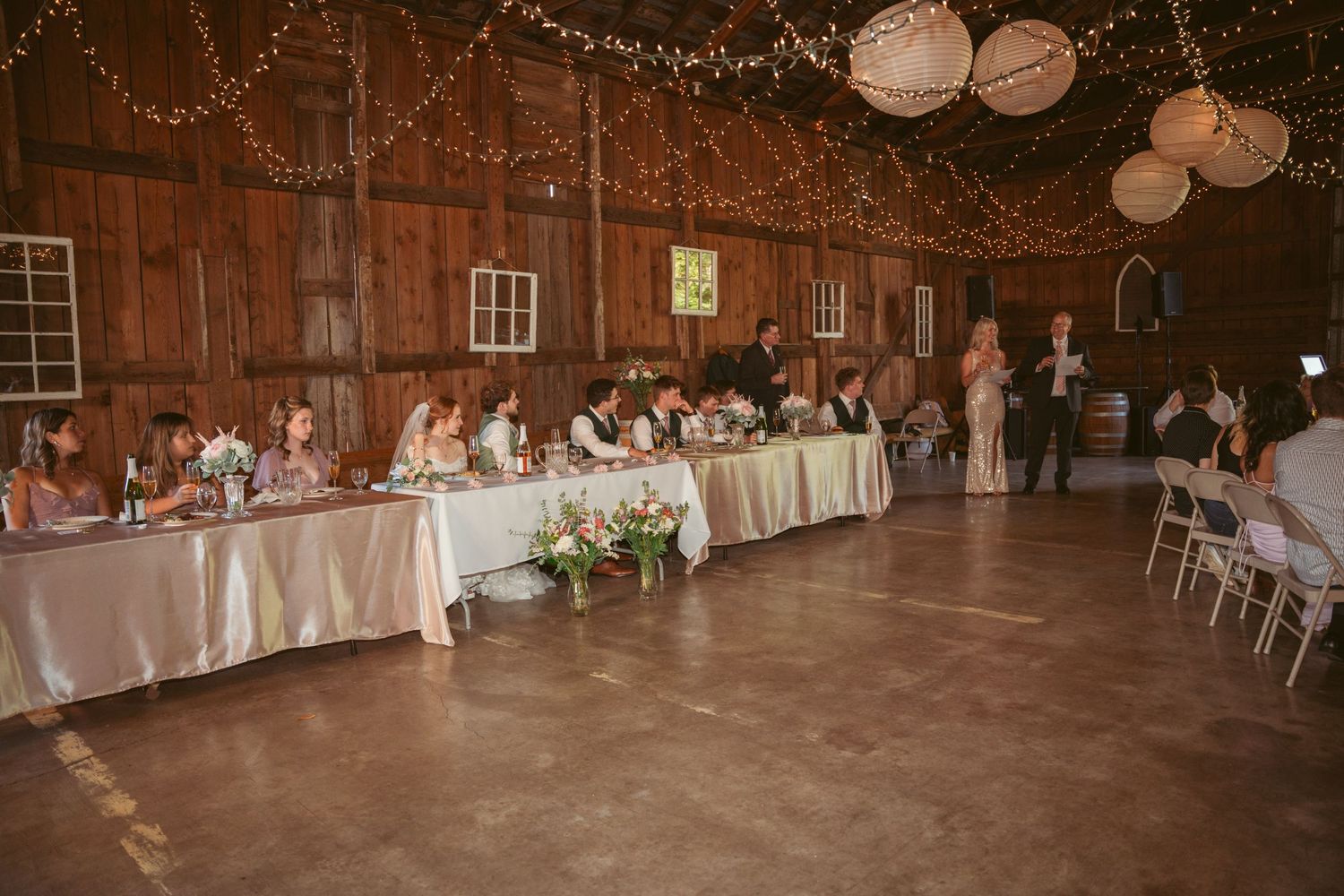 Wedding reception setup in rustic barn with string lights and white tablecloths.