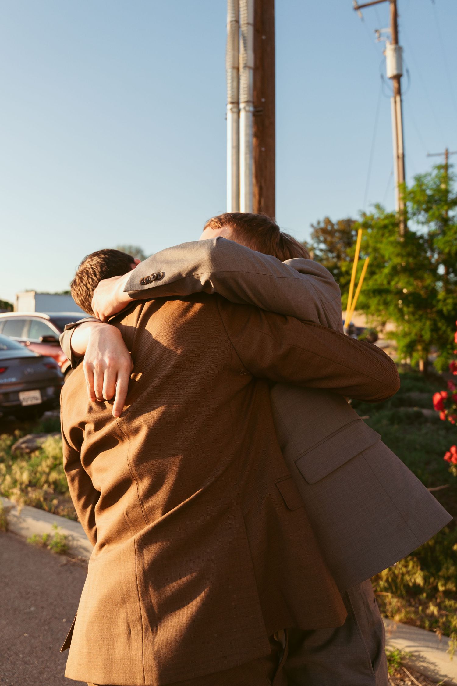 Two people share an emotional embrace outdoors in brown formal attire.