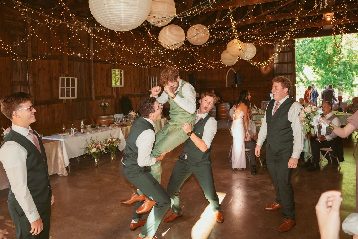 Wedding party performs choreographed dance moves in a barn with twinkling lights and lanterns.