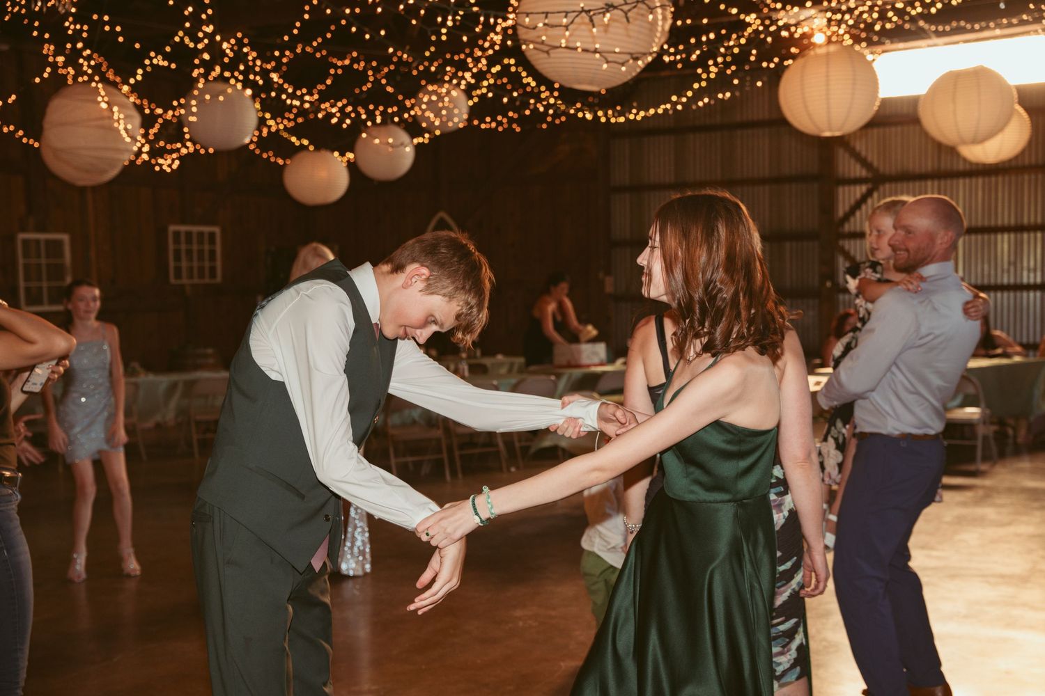 Couples swing dancing on a wooden floor under warm string lights at a barn reception.