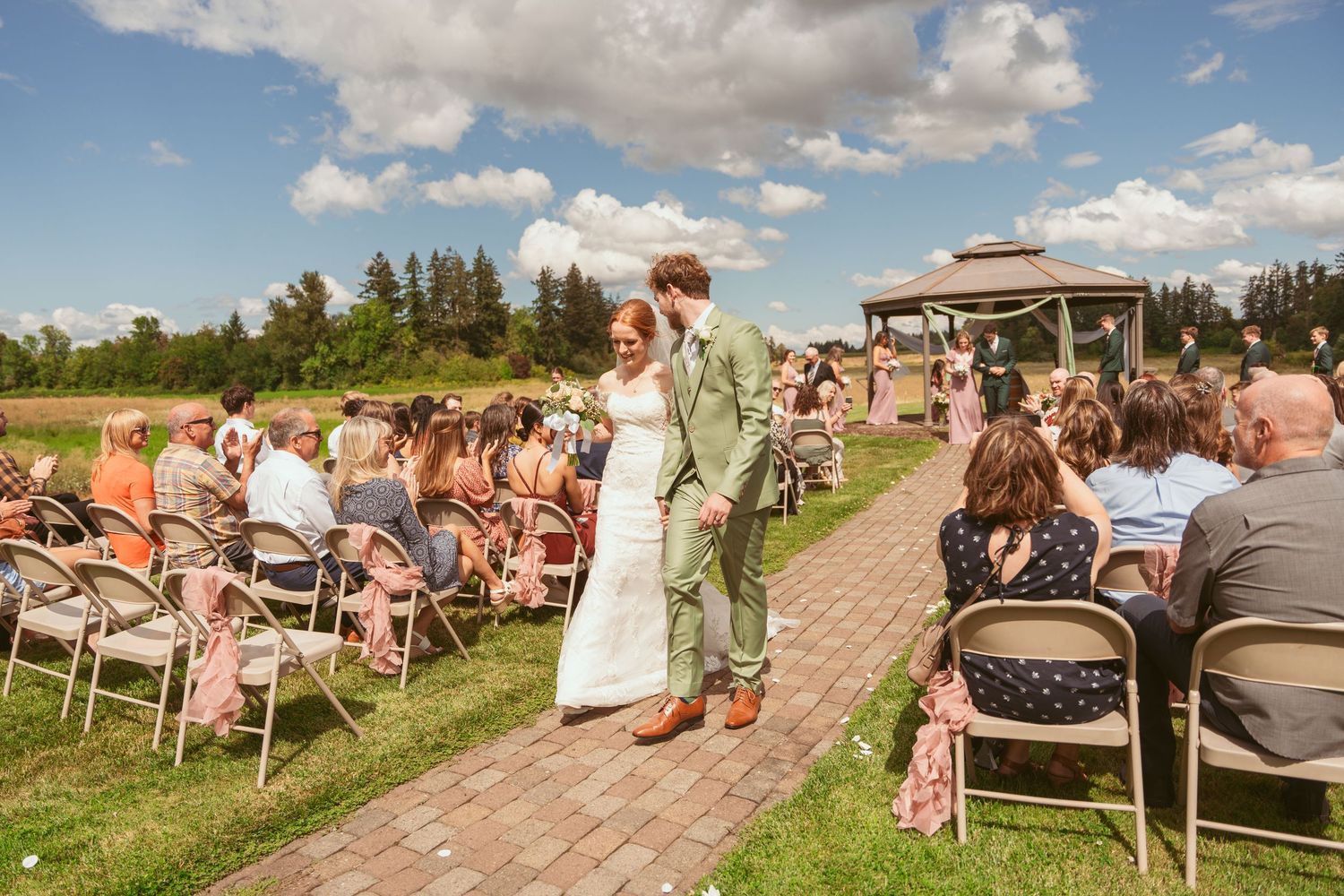 Outdoor wedding ceremony with guests seated on white chairs under a cloudy sky.