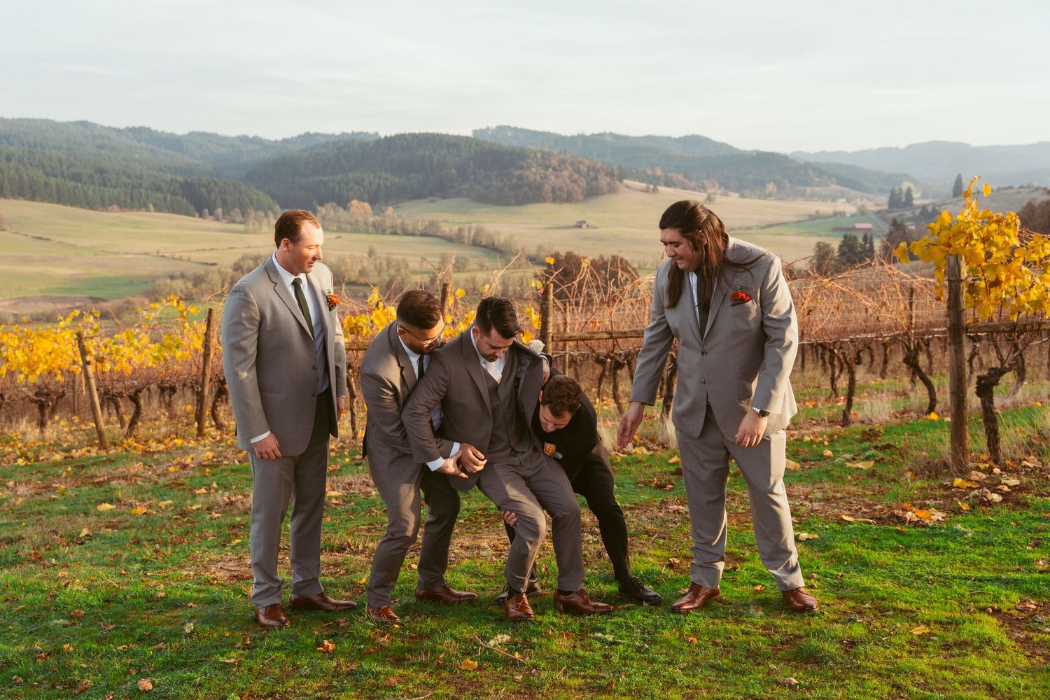 Groomsmen in matching suits share a lighthearted moment on a hillside vineyard.