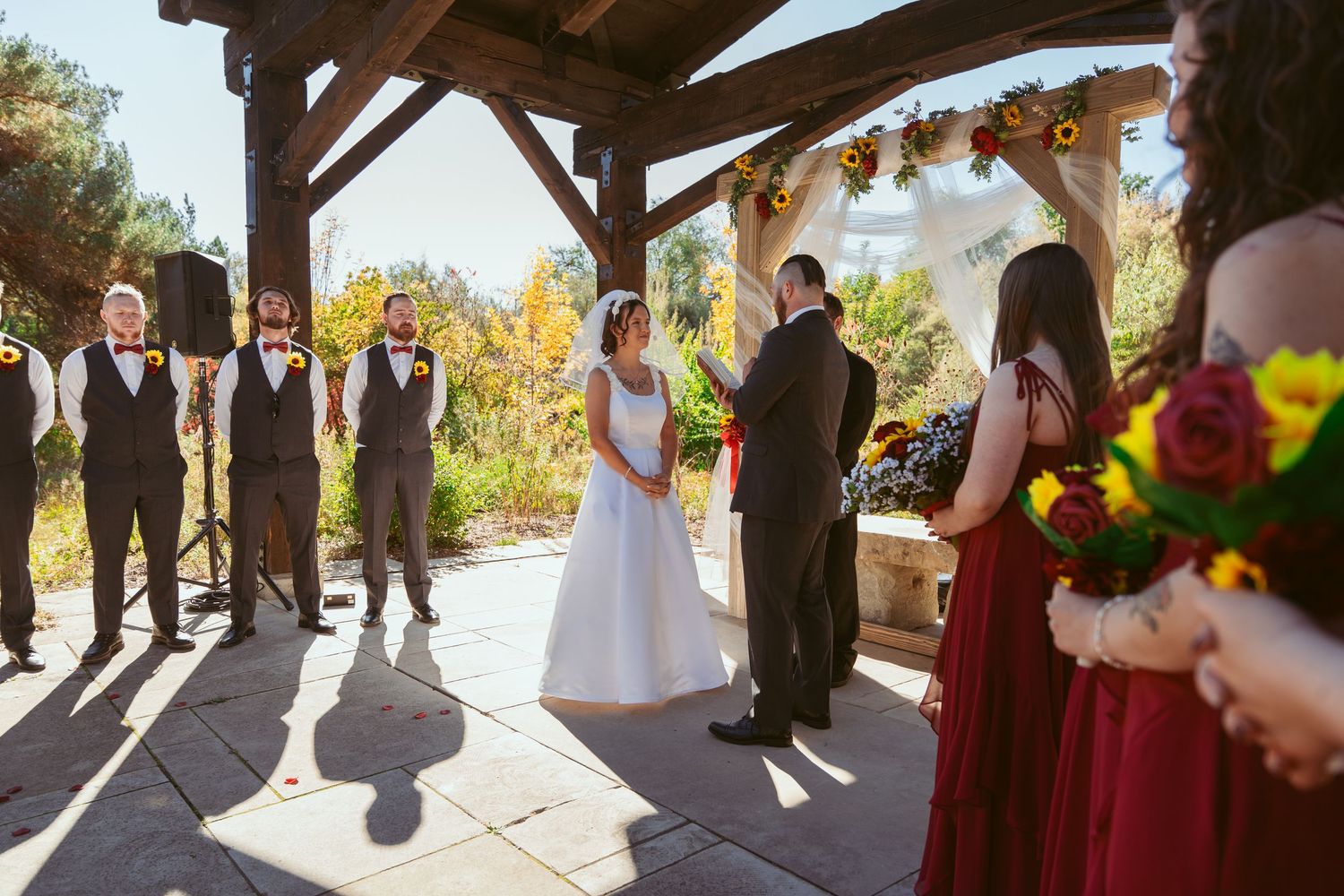 Wedding party standing in silhouette under wooden pavilion with red and yellow floral accents.