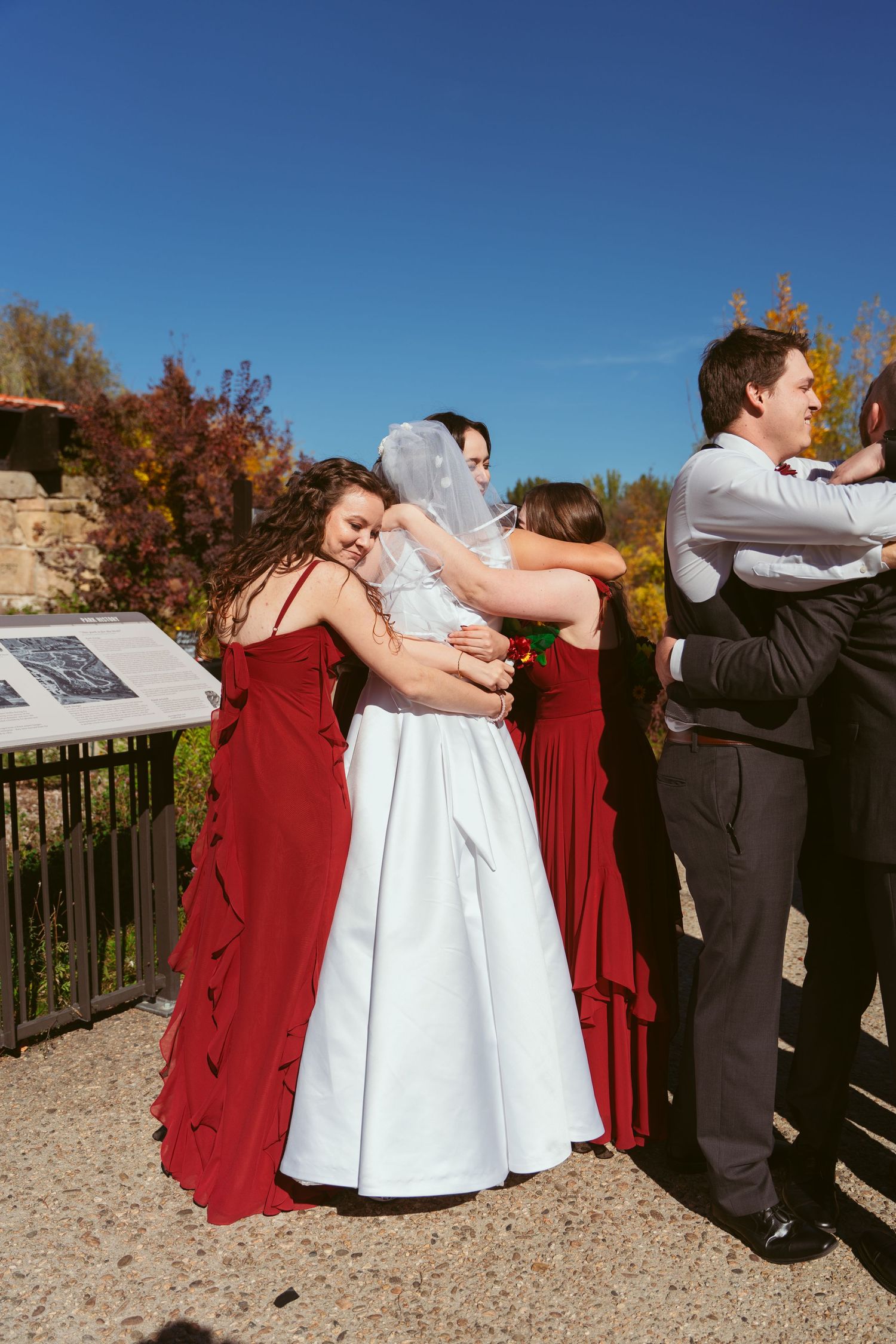 Wedding party in red and black attire poses playfully on a gravel path during an autumn outdoor photoshoot.