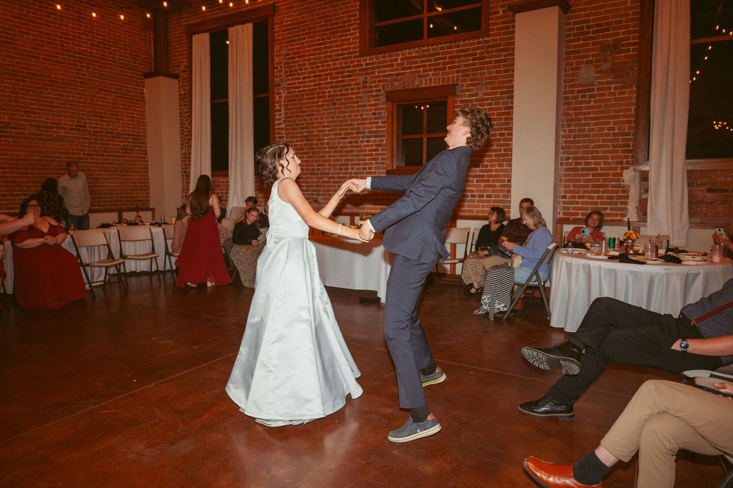 Guests enjoying a dance at a wedding reception in a brick-walled venue with overhead lighting.