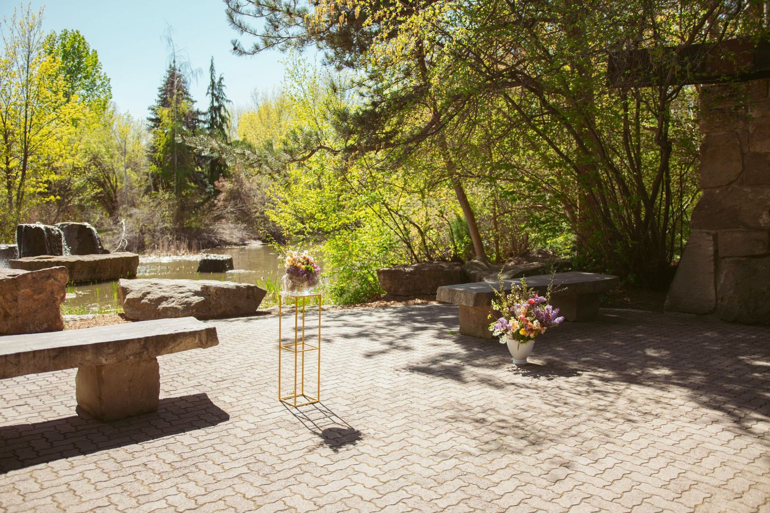 A sunny brick patio with stone benches, decorative planters and lush greenery in the background on a spring day.