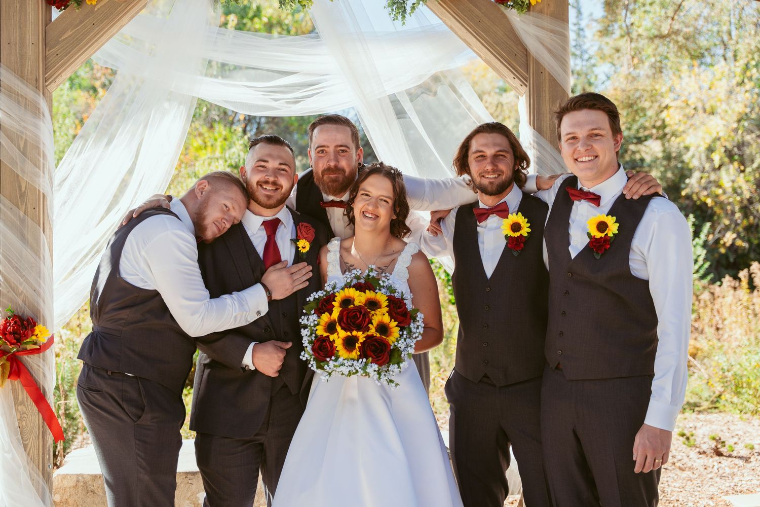Wedding group stands together under rustic pavilion decorated with sunflowers and white draping.