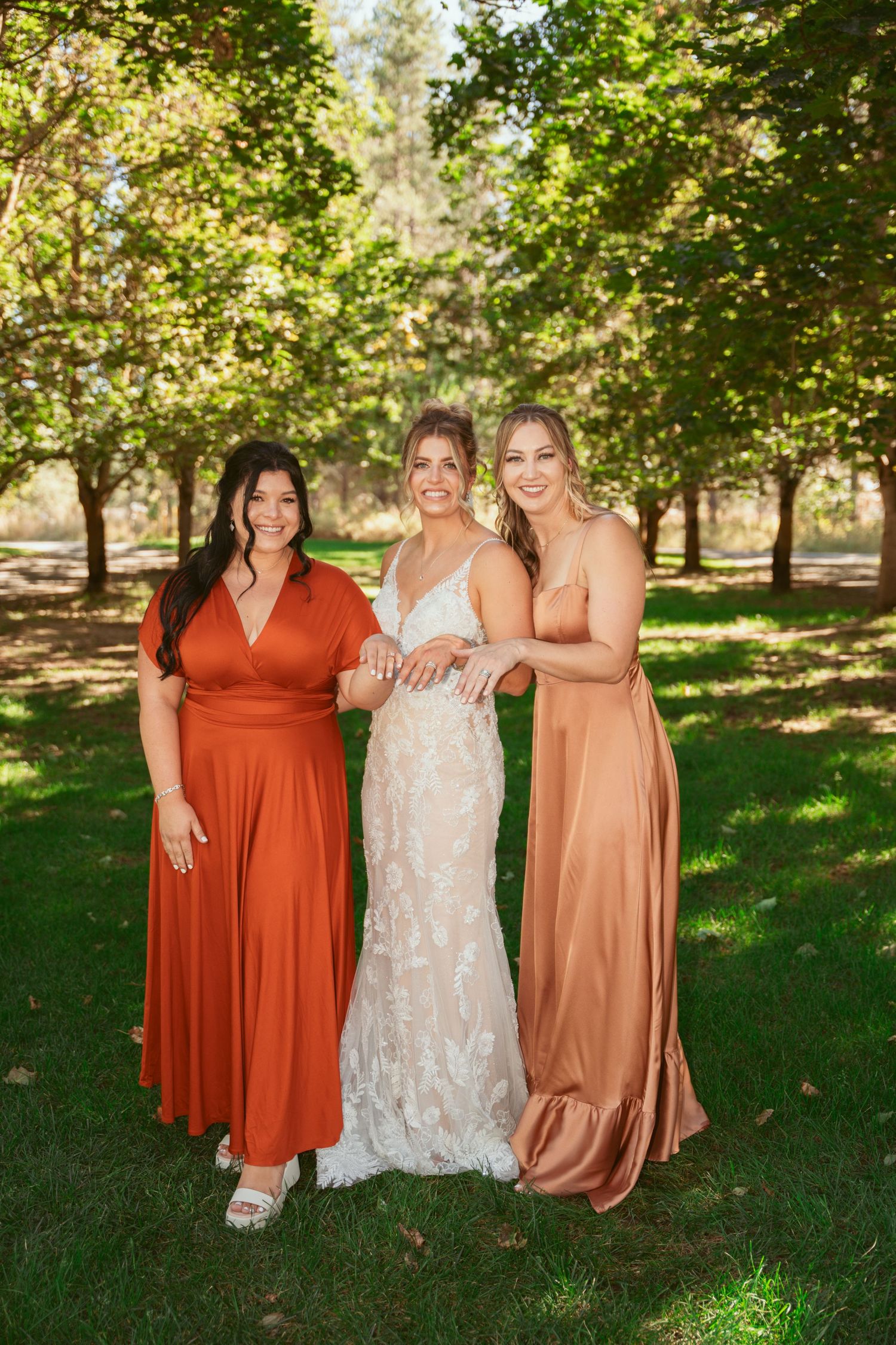 Three women in elegant dresses pose together outdoors in a sunlit garden setting with trees in the background.