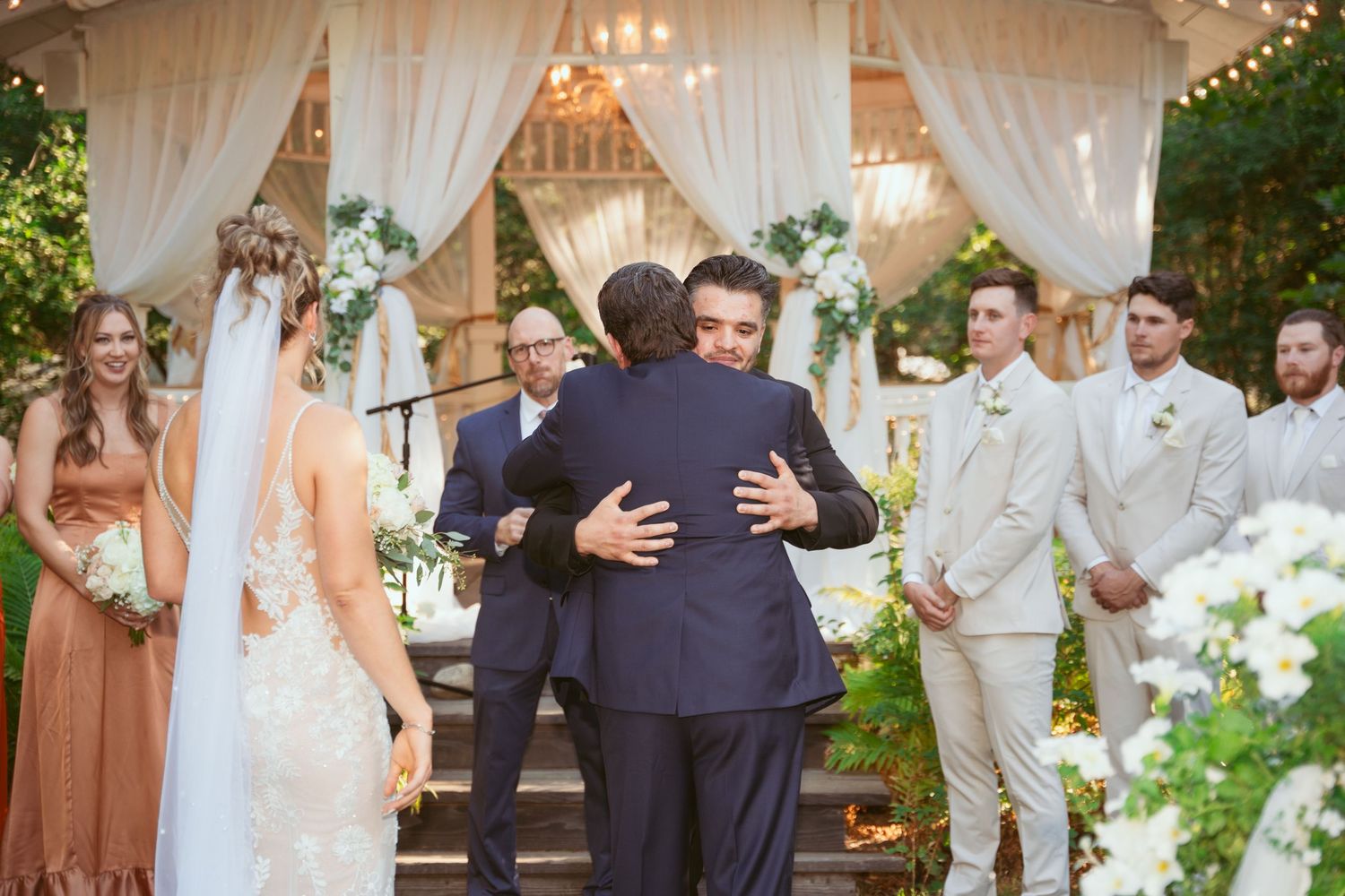 Wedding ceremony under an elegant white draped gazebo with floral decorations and guests seated.