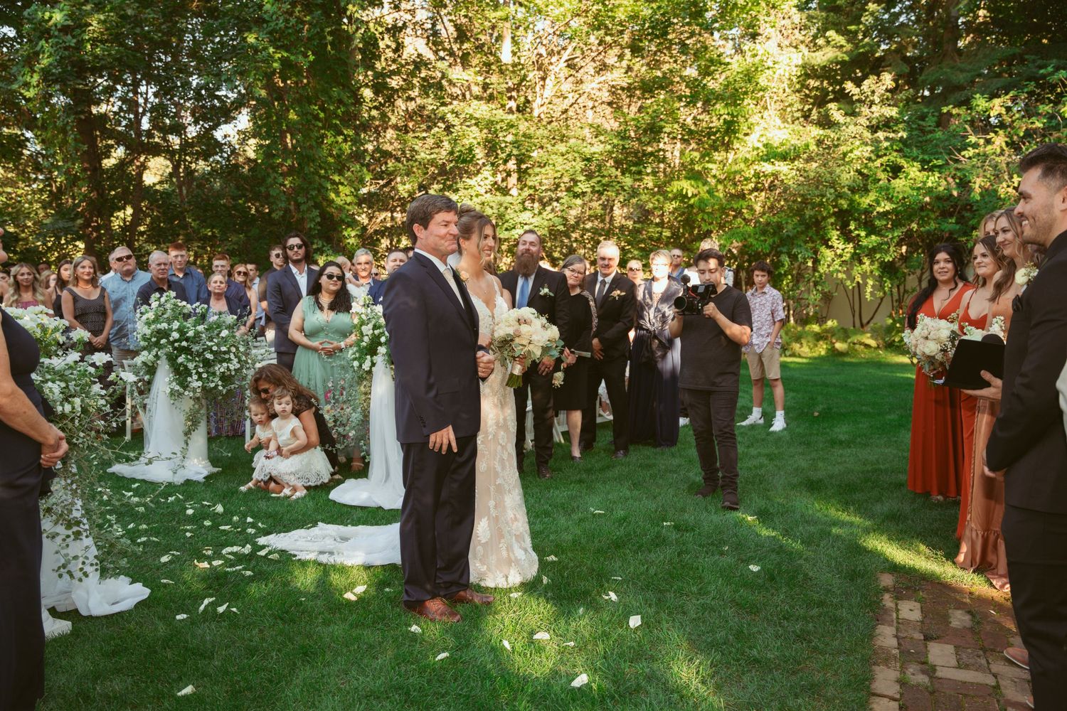 An outdoor garden wedding ceremony with guests gathered on a lush green lawn surrounded by trees and scattered flower petals.