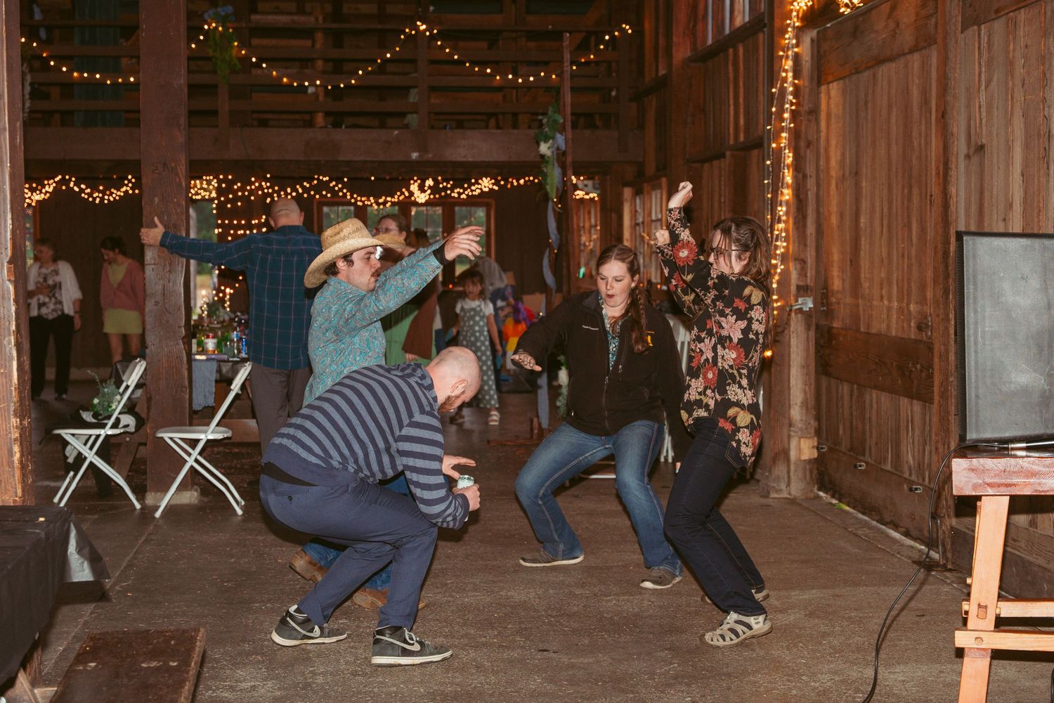 Guests dancing together in a rustic wooden barn decorated with string lights during a celebration.