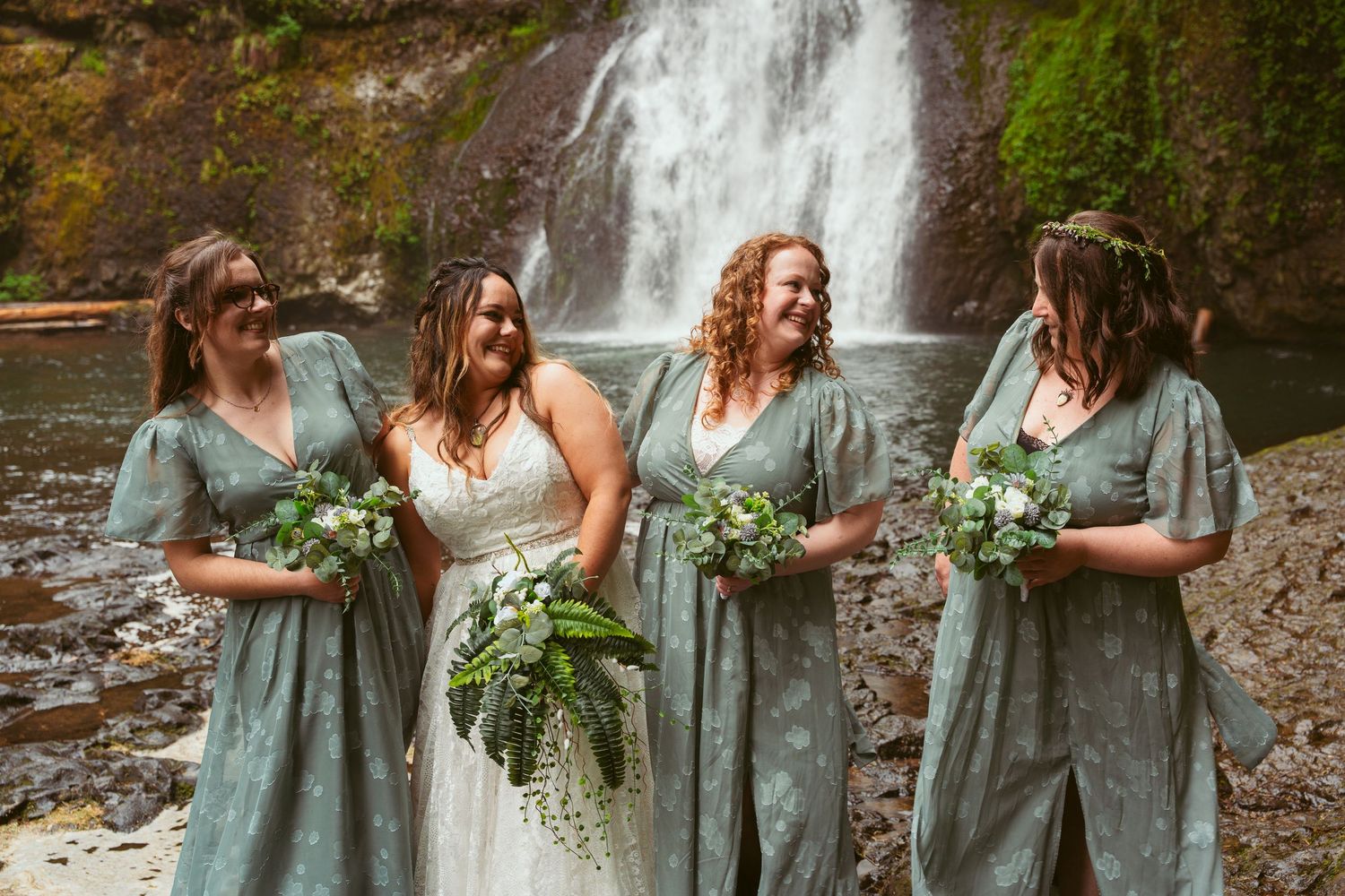 Bridesmaids in sage green dresses share candid moments by a forest waterfall.