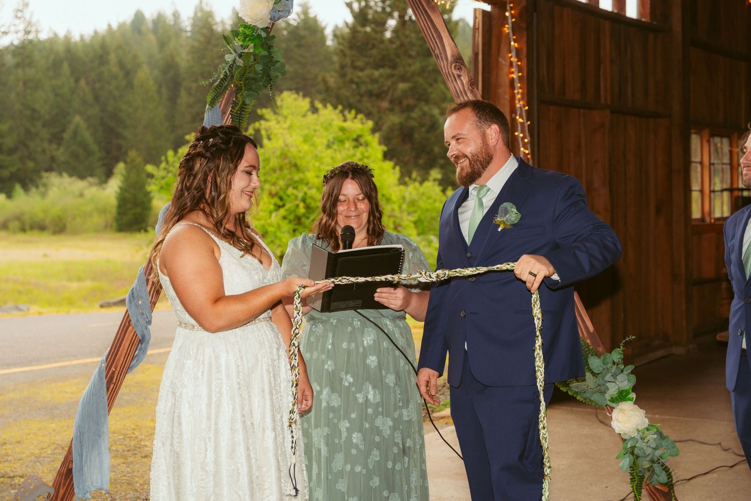 Wedding party poses inside rustic barn with forest visible through open doorway.