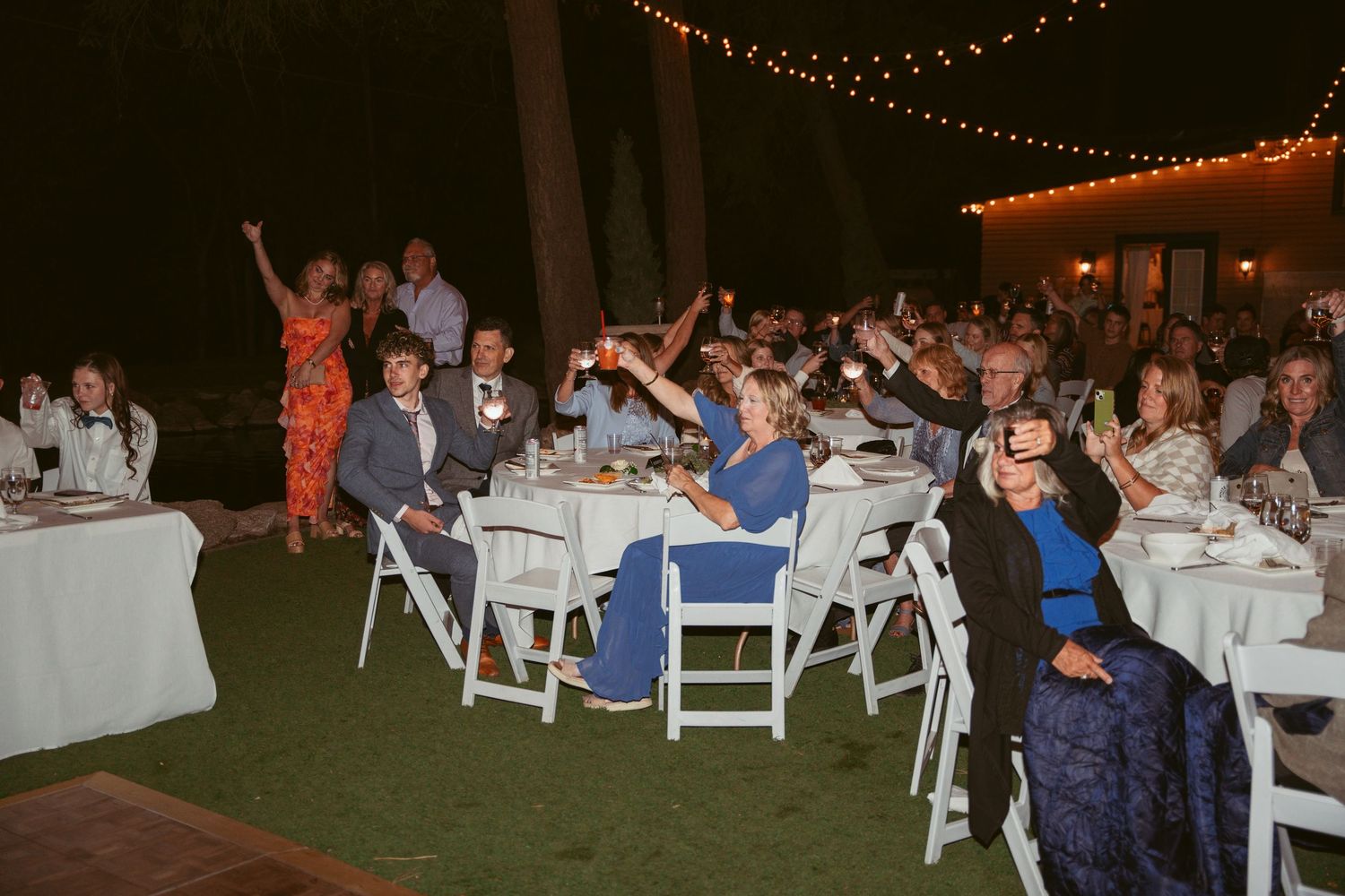 Wedding guests dance and celebrate under twinkling string lights at an outdoor evening reception.