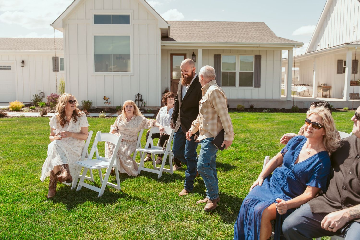 A group gathers outside a white farmhouse-style building on a sunny day.