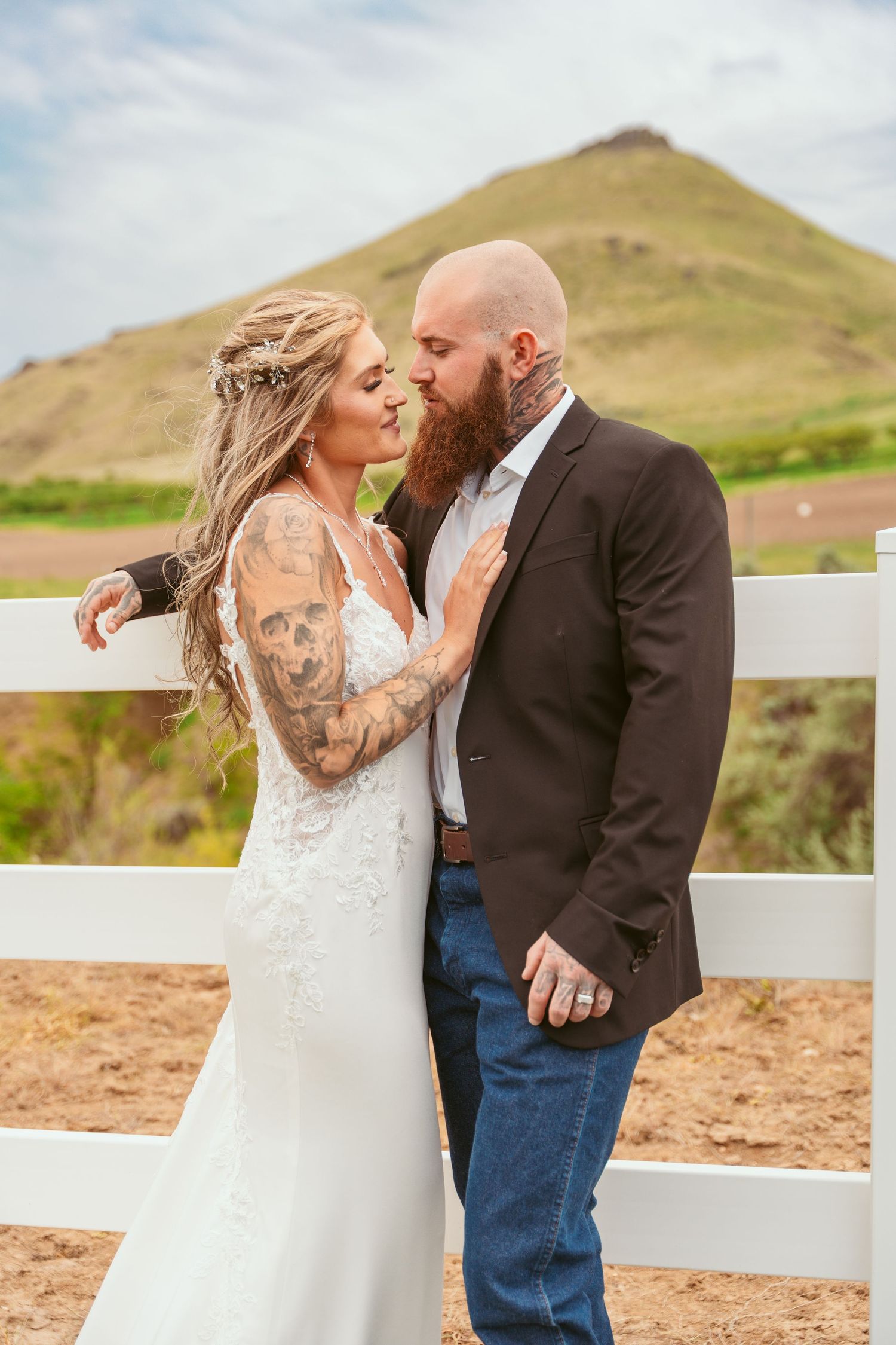 Couple shares intimate moment against rustic white fence and mountain backdrop.