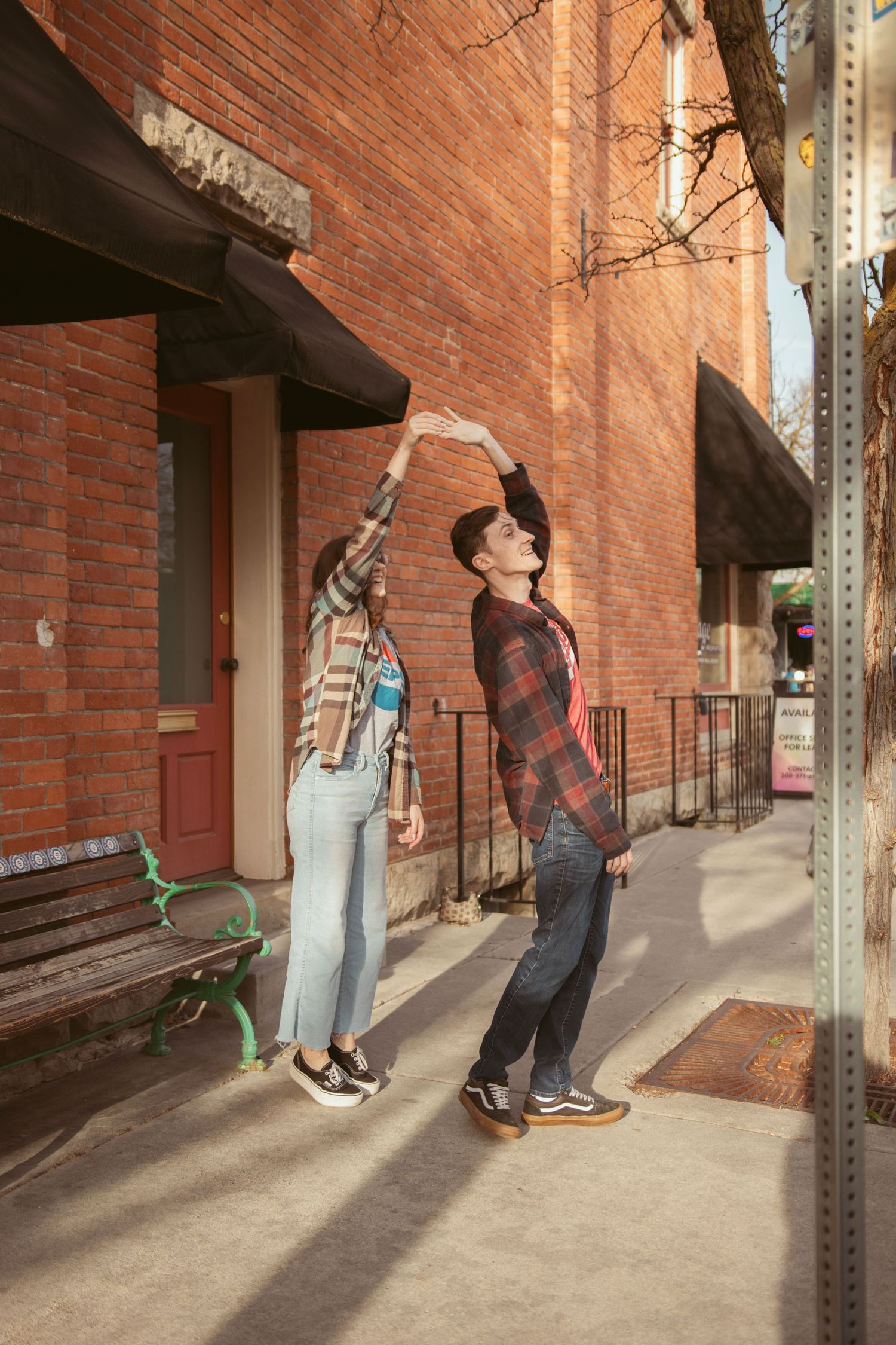 Two people stretch their arms upward while posing against a red brick building exterior.