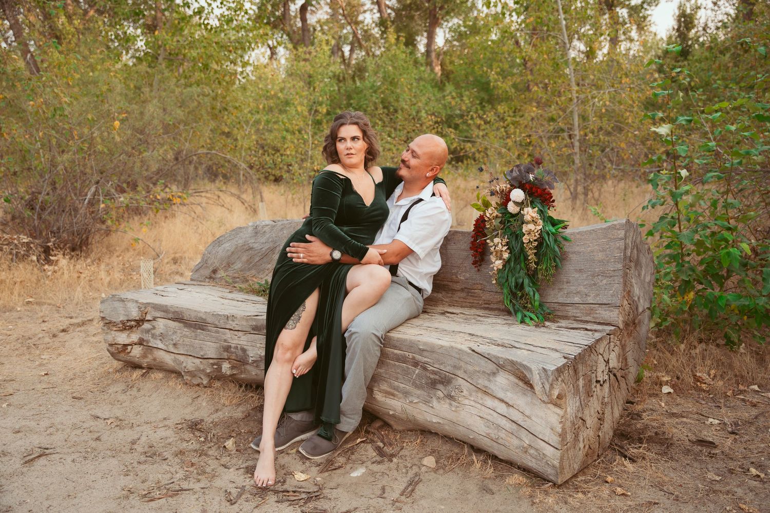 A couple sharing intimate moments while seated on a fallen log in a wooded area.