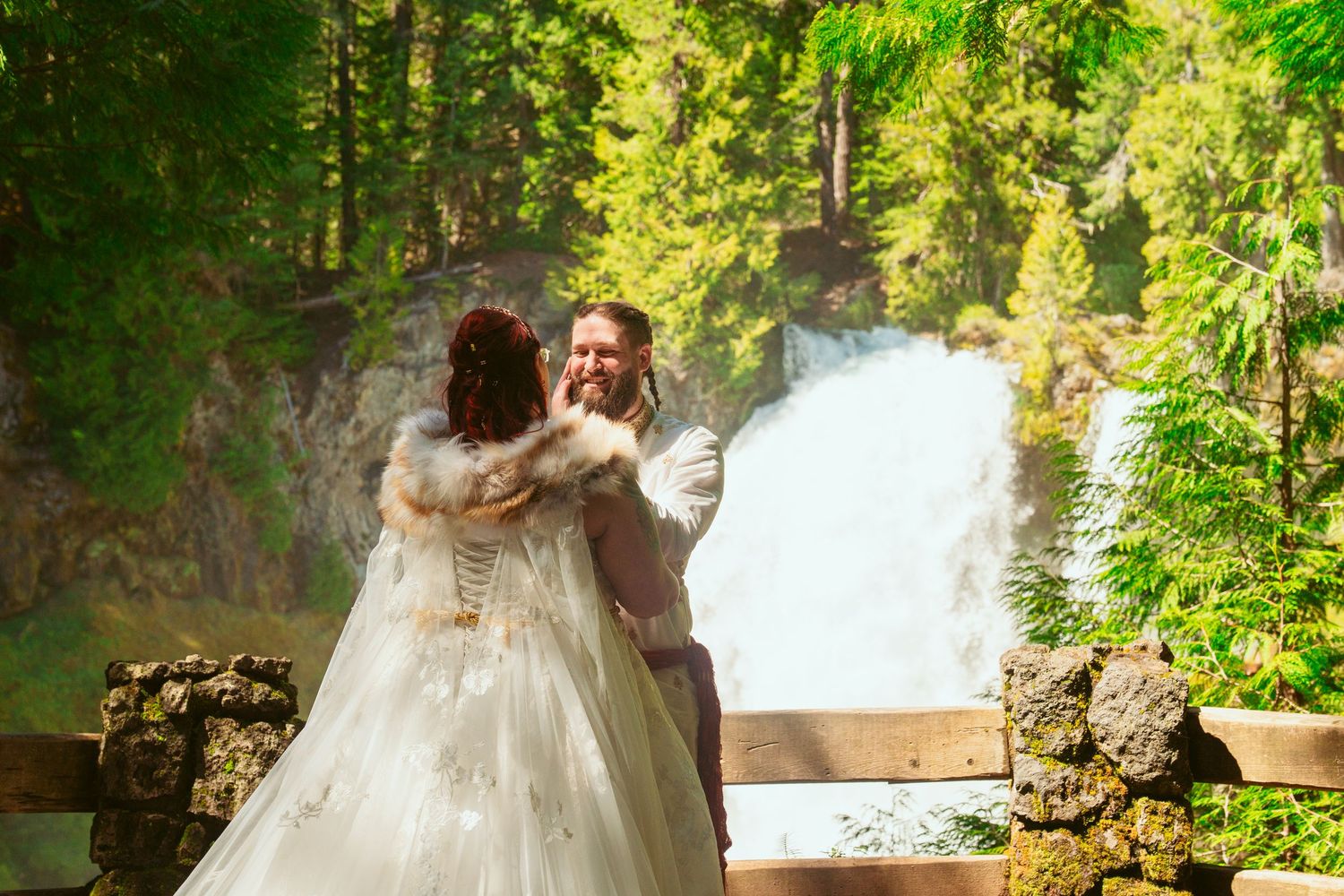 A couple in white wedding attire dances together near a scenic waterfall.