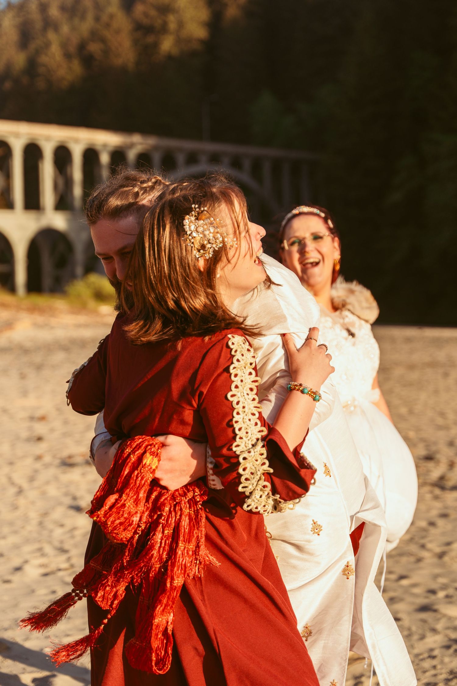 A couple in red and white outfits dance joyfully on a beach with cliffs and trees in the background at sunset.