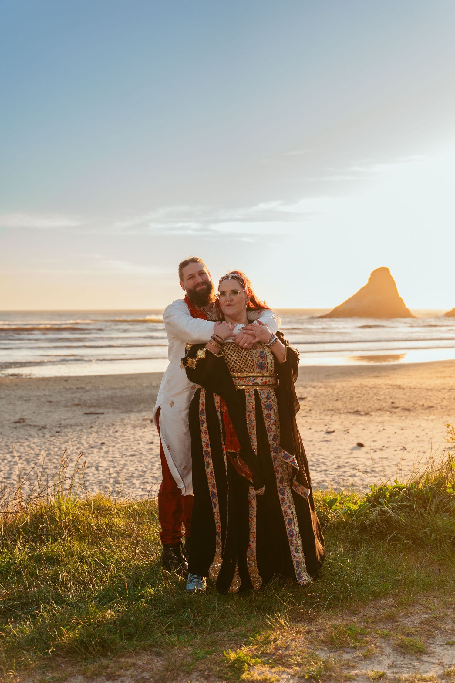 A couple in fantasy-themed wedding attire embracing on a scenic beach at golden hour.