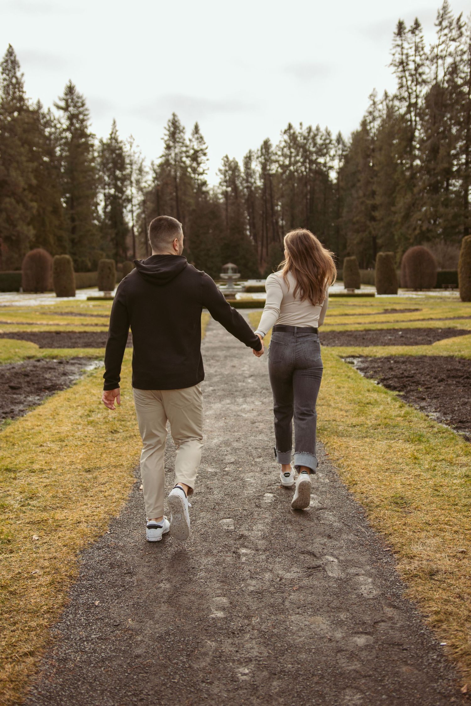 Couple walking hand in hand down a path surrounded by evergreen trees and yellow fields.