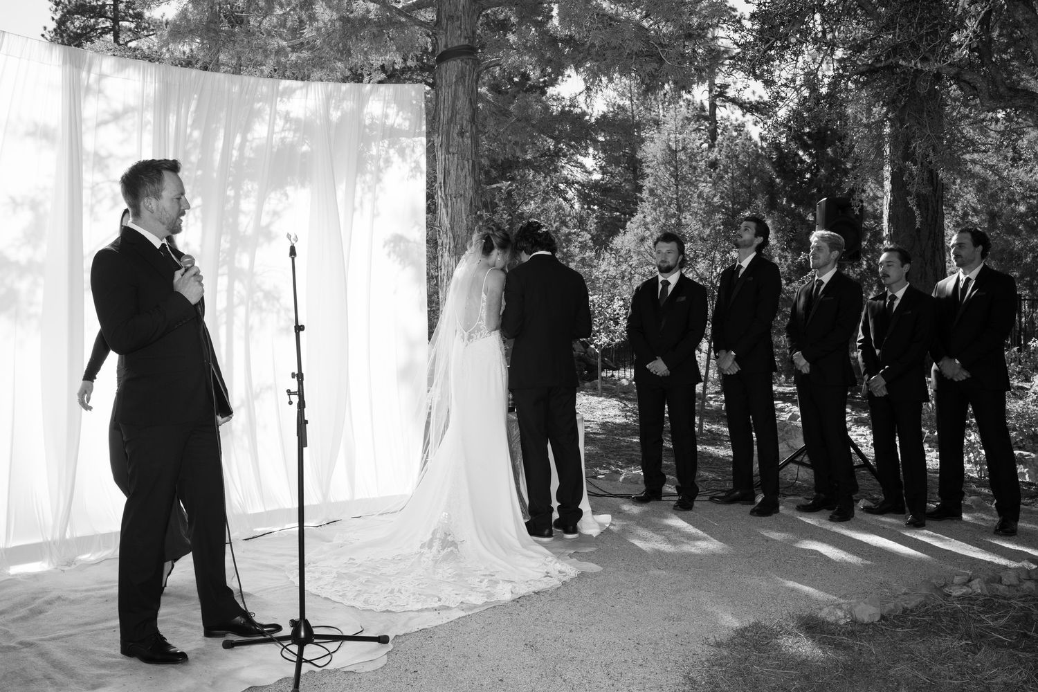A black and white photo of a wedding ceremony with attendants lined up beside white draping outdoors.