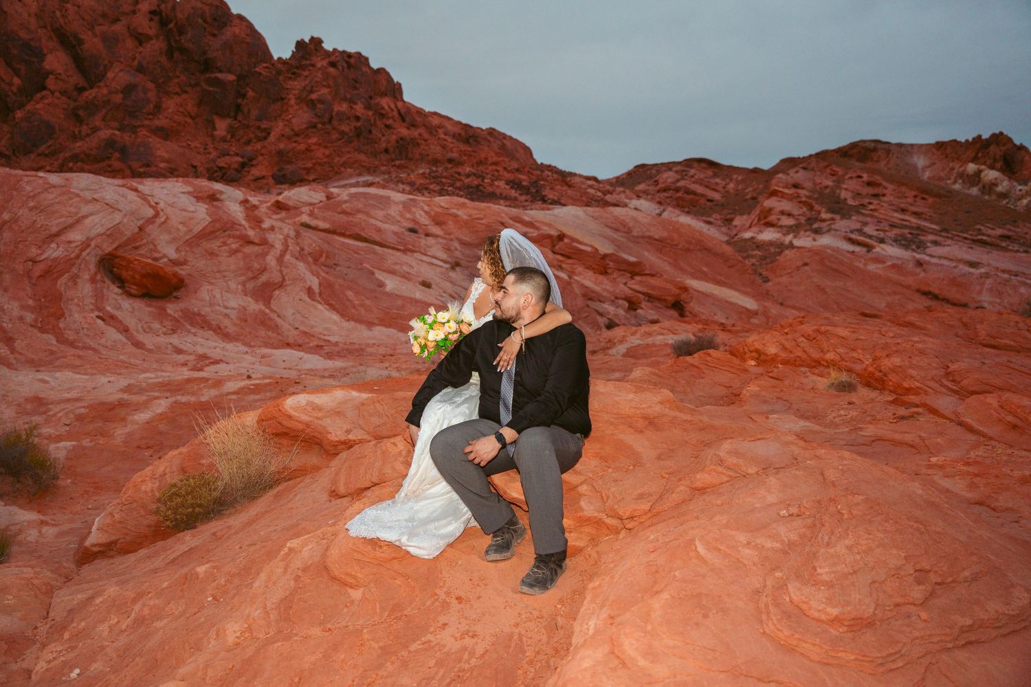 Couple posing on red sandstone cliffs with cloudy sky backdrop during sunset.