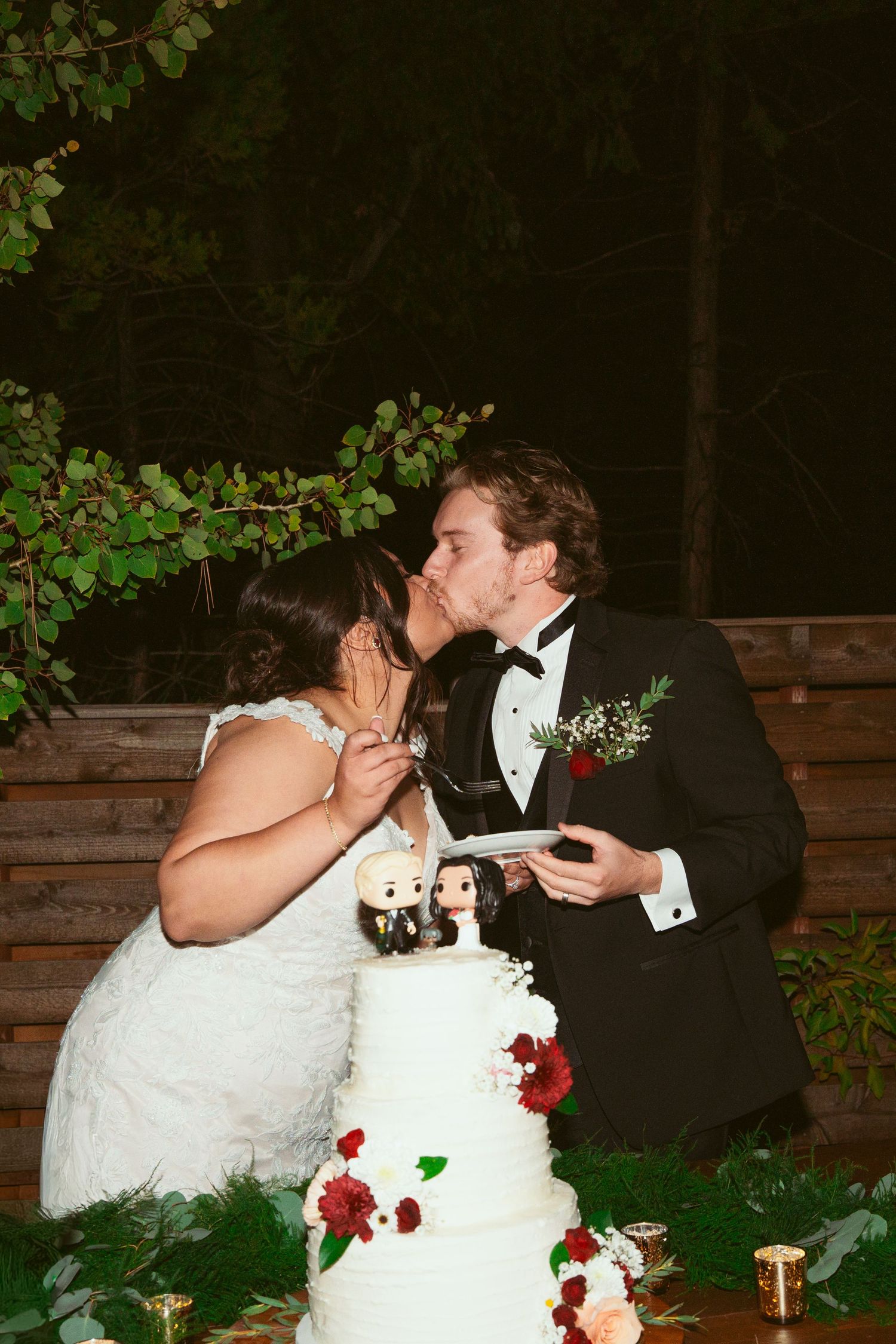 Newlyweds share cake cutting moment beside elegant white wedding cake decorated with red roses.