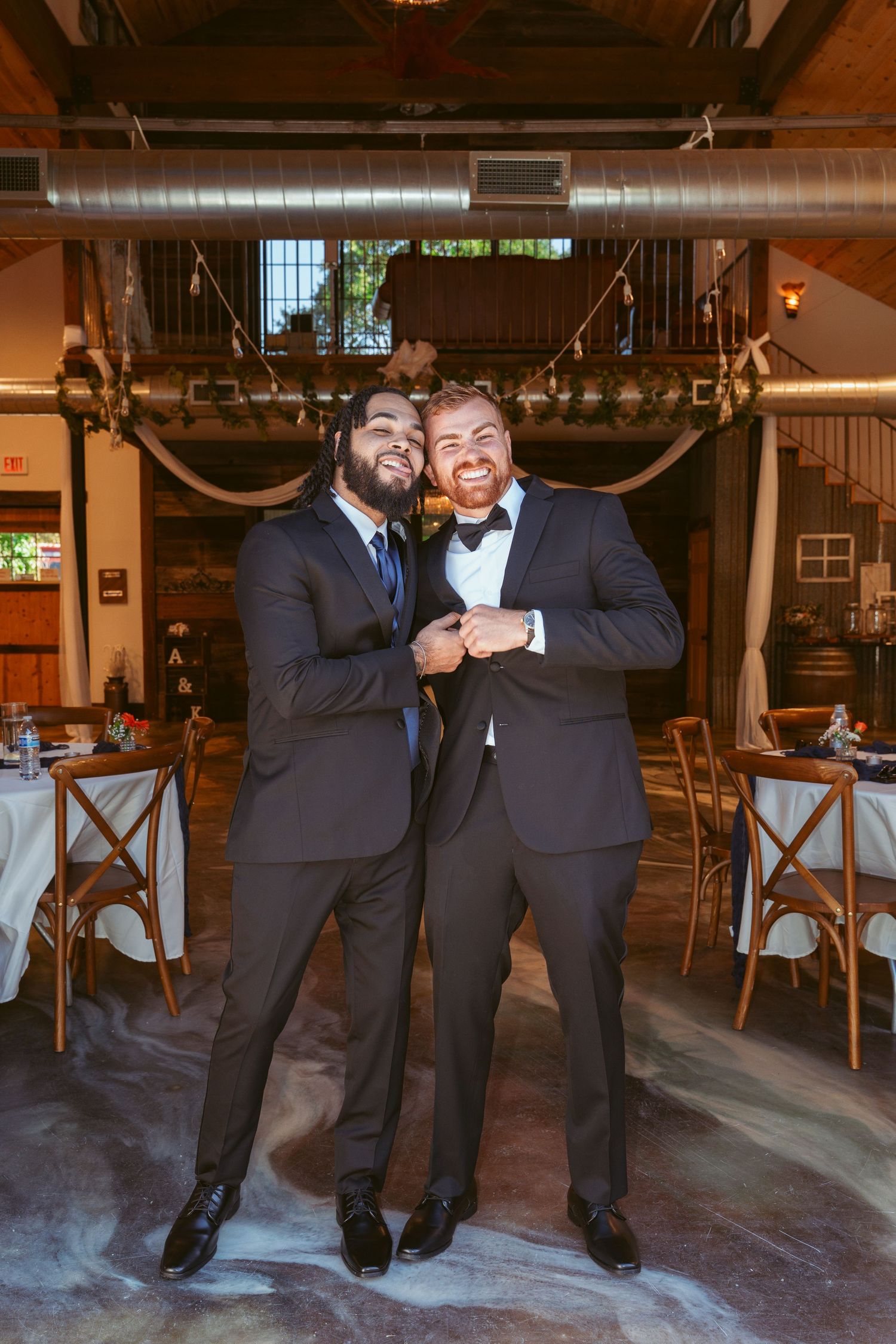 Two men in formal suits sharing a joyful moment at an indoor wedding venue.