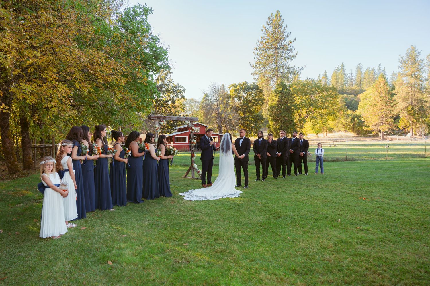 Wedding celebration group photo on expansive lawn with autumn trees.