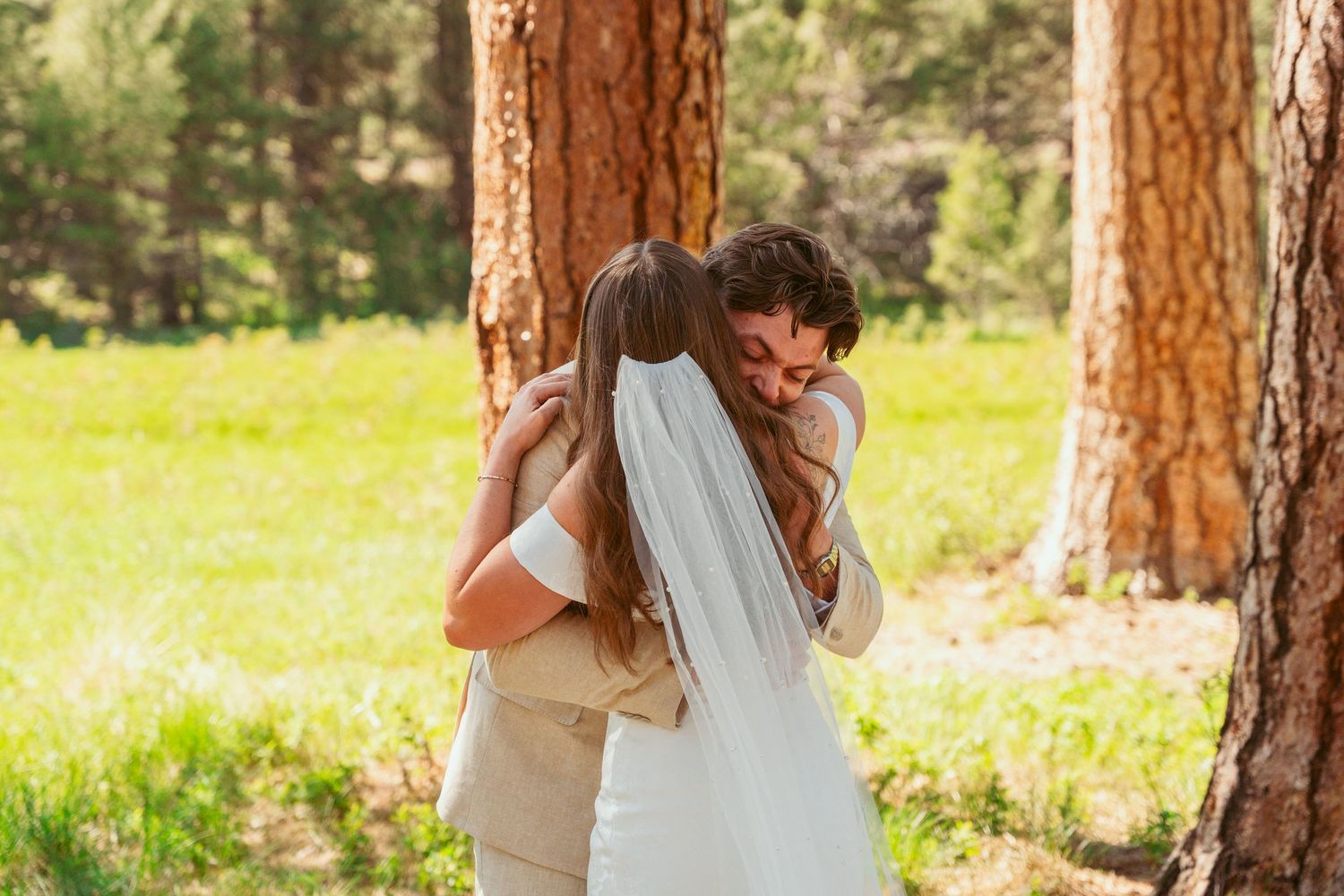 A wedding couple shares a playful first look moment among tall pine trees in a sunny meadow.