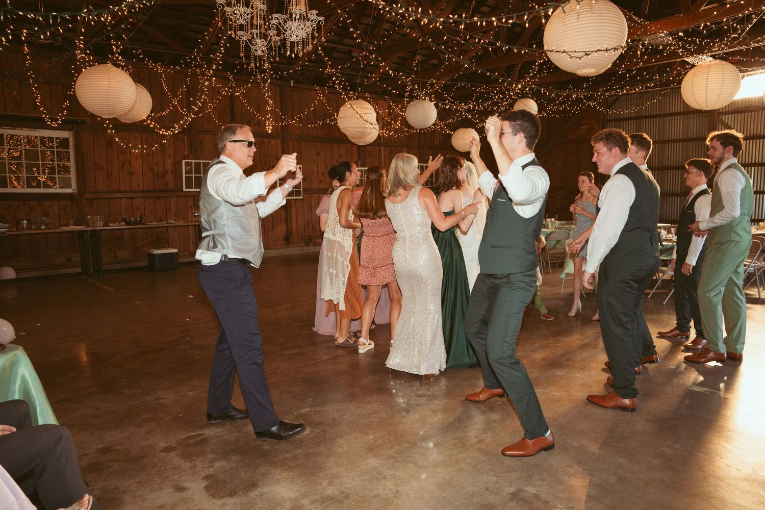Wedding party members dance together under warm lighting and paper lanterns at an indoor reception.