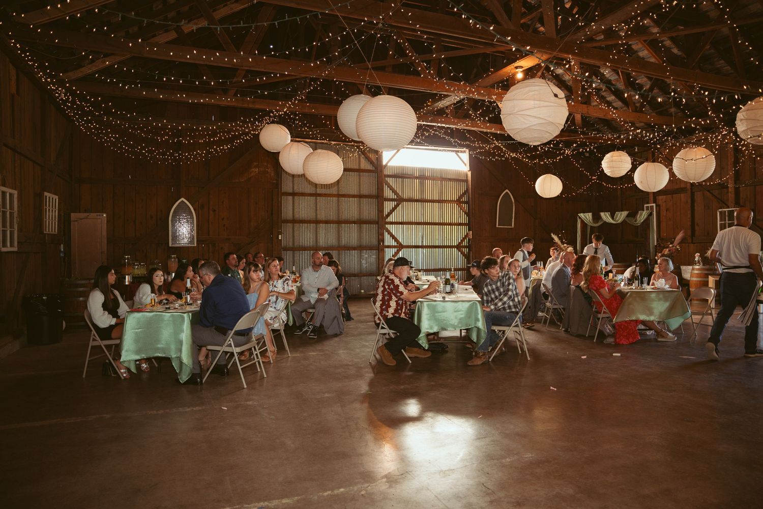 Evening wedding celebration in a barn with ambient lighting and casual seating arrangements.
