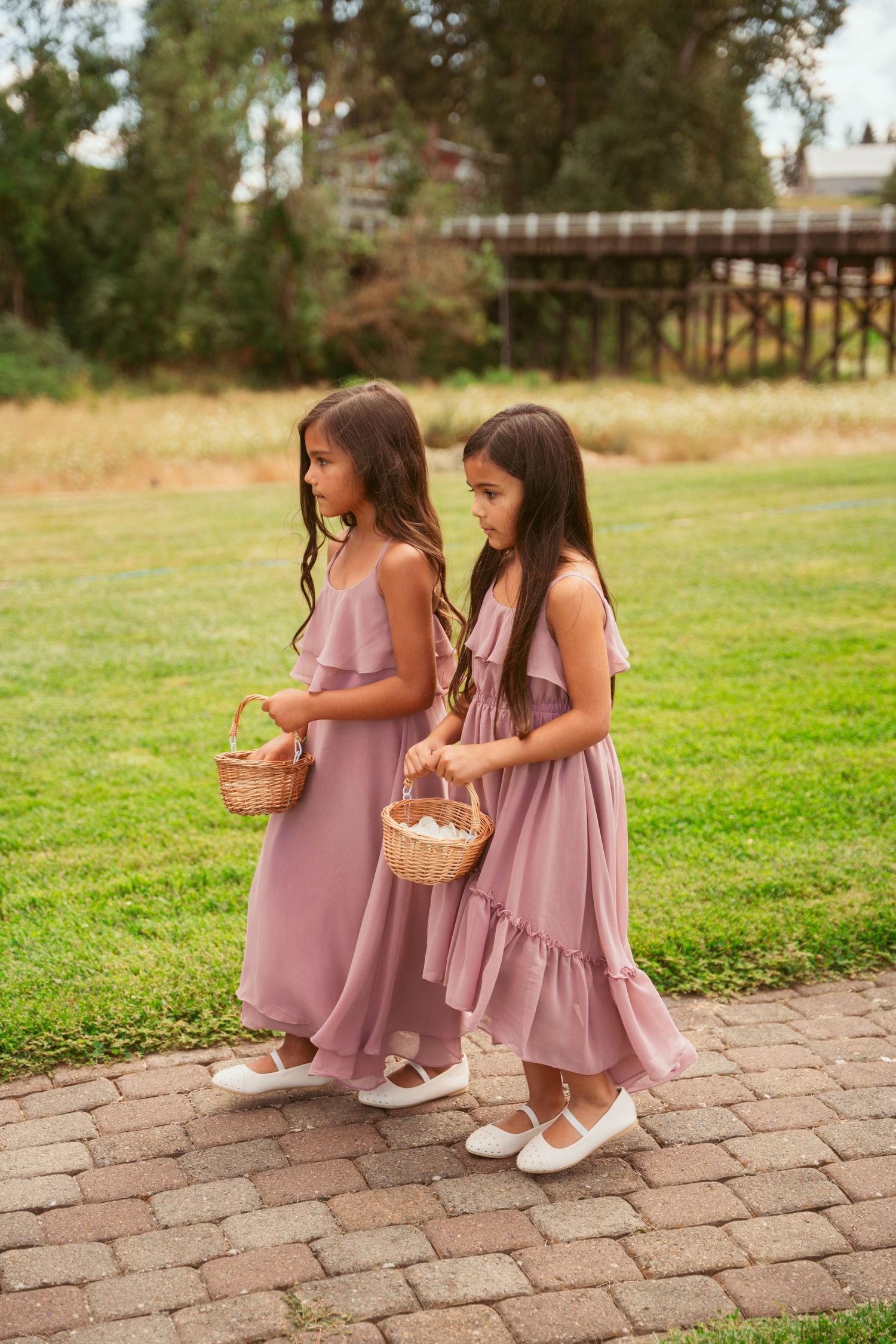Children in matching mauve dresses carry flower baskets down a brick pathway.