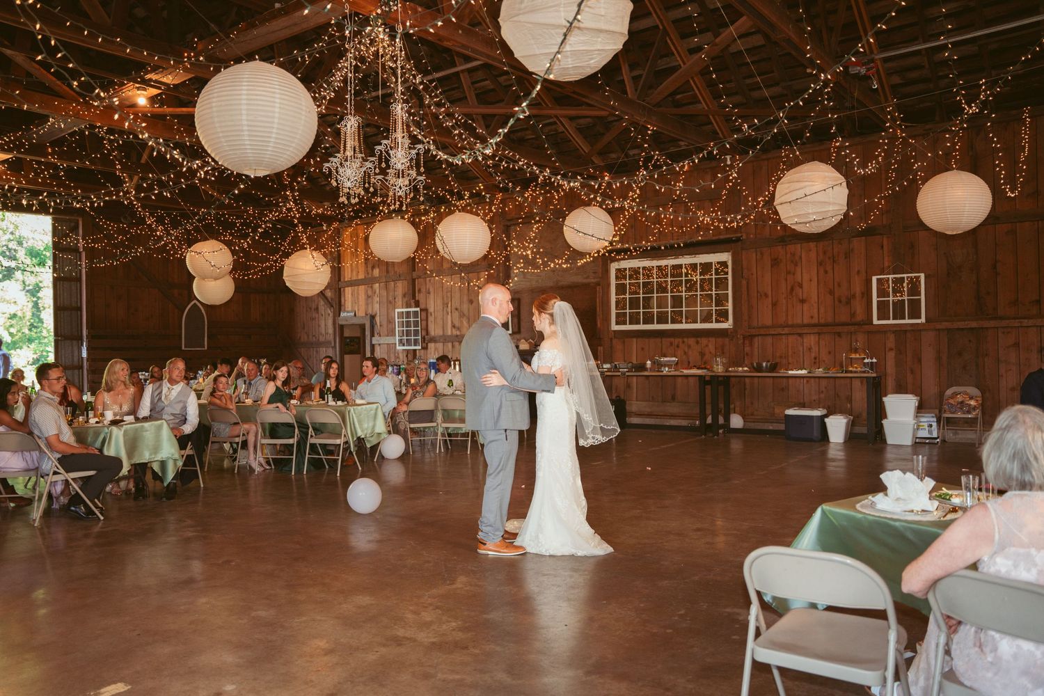 Wide view of a barn wedding reception with white paper lanterns and string light decorations.