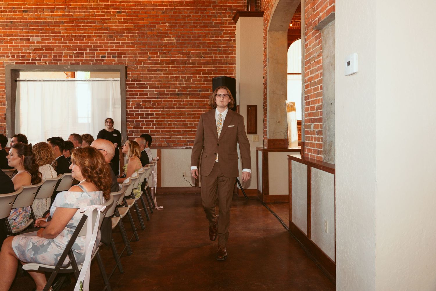 Wedding ceremony processional in rustic brick venue with guests seated along aisle.