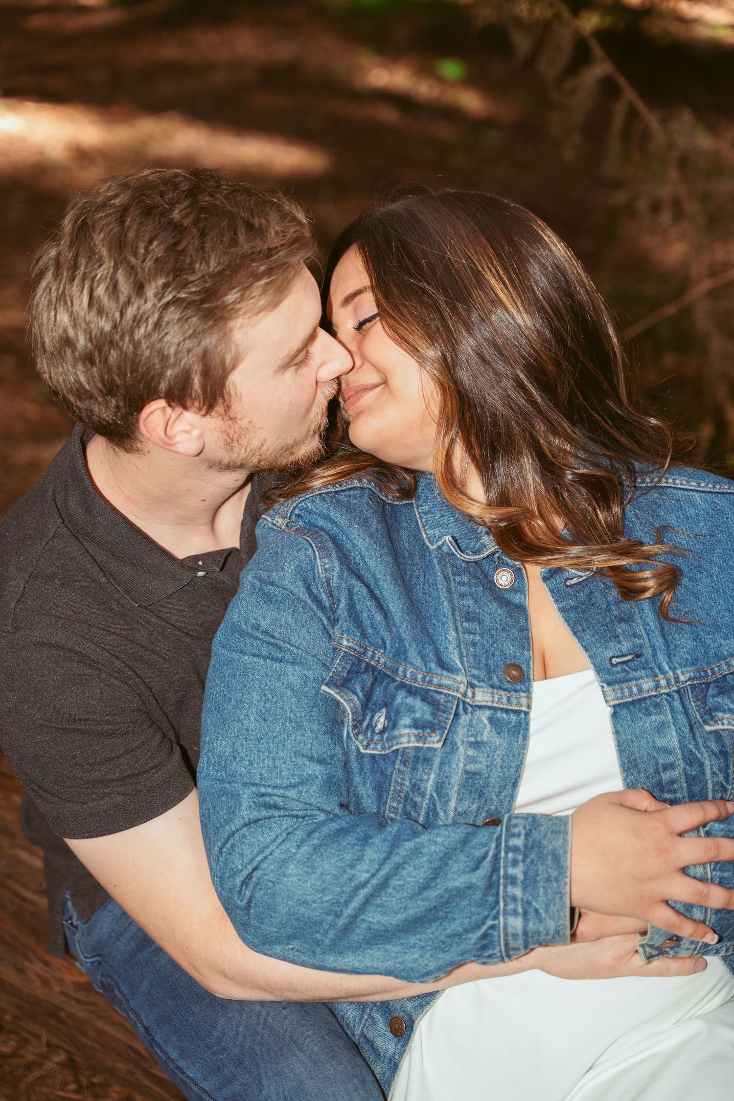 A playful couple shares laughter and kisses while sitting in a redwood forest.
