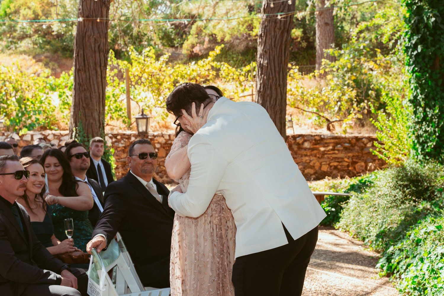 An emotional wedding ceremony moment under tall trees with guests looking on.