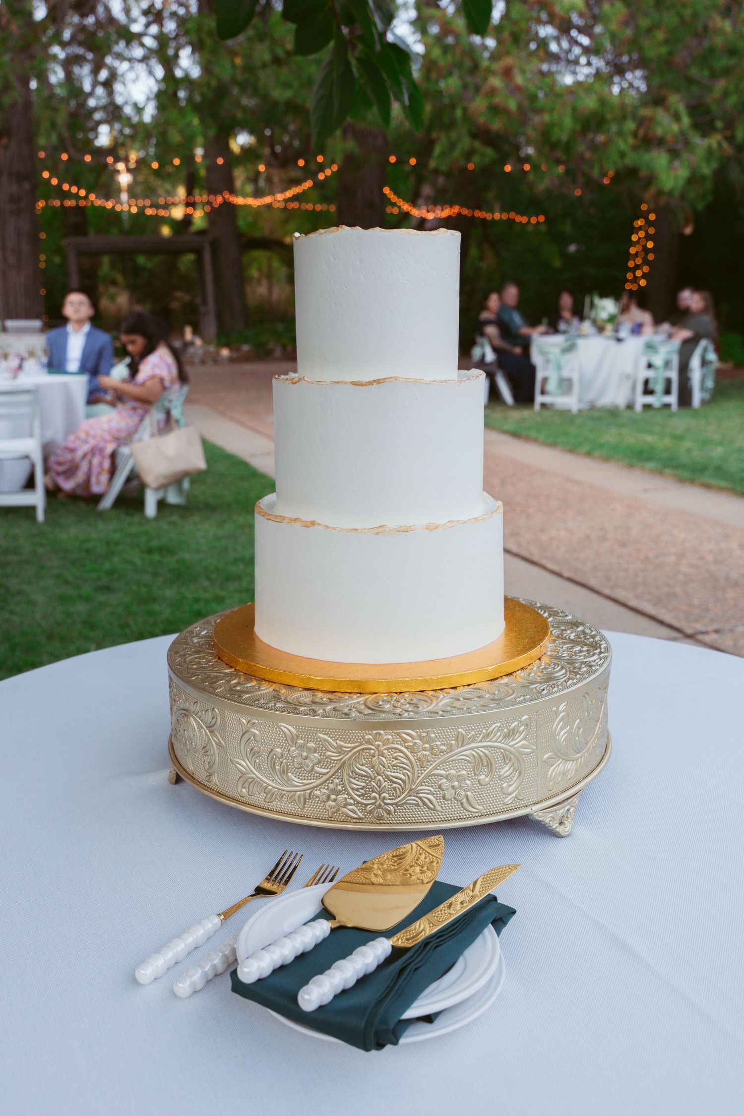 Three-tiered white wedding cake with gold trim displayed outdoors under string lights.