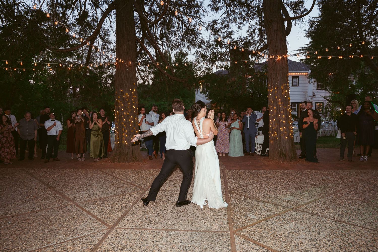 Wedding guests gather for dancing on an outdoor dance floor surrounded by trees and twinkle lights.
