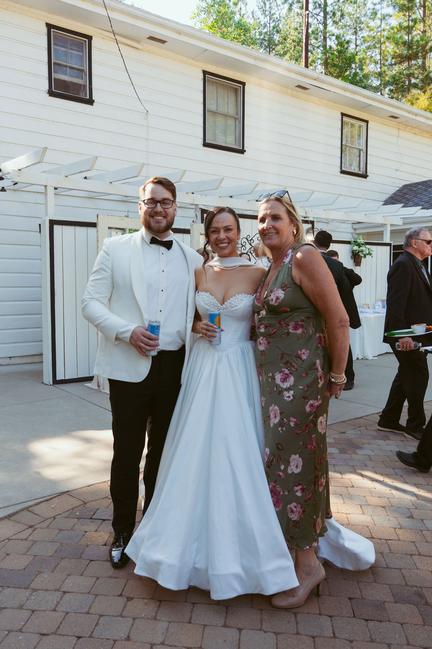 Wedding party members pose together on brick patio outside white building during daytime celebration.