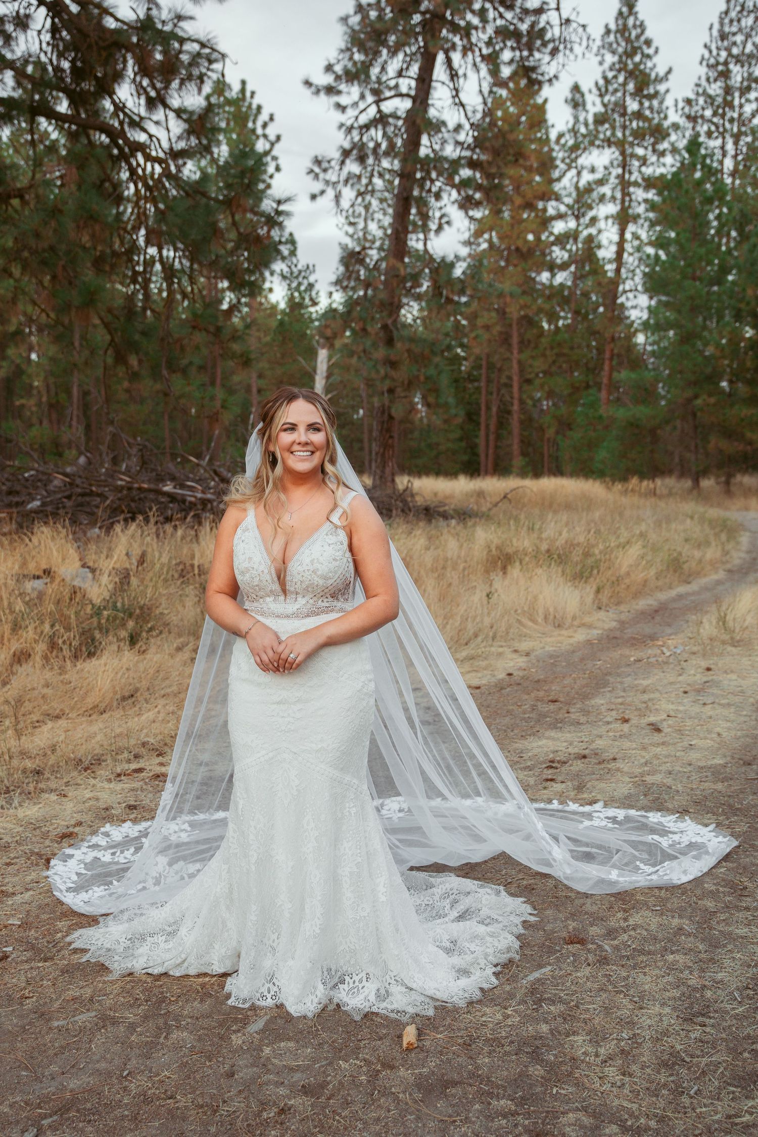 Bride in lace gown with cathedral veil poses on forest path surrounded by pine trees.